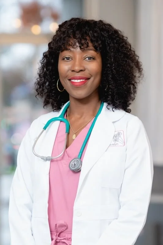 A female doctor or medical professional smiling, wearing a white coat and a stethoscope around her neck, with curly black hair and gold hoop earrings, standing in a clinical or hospital setting.