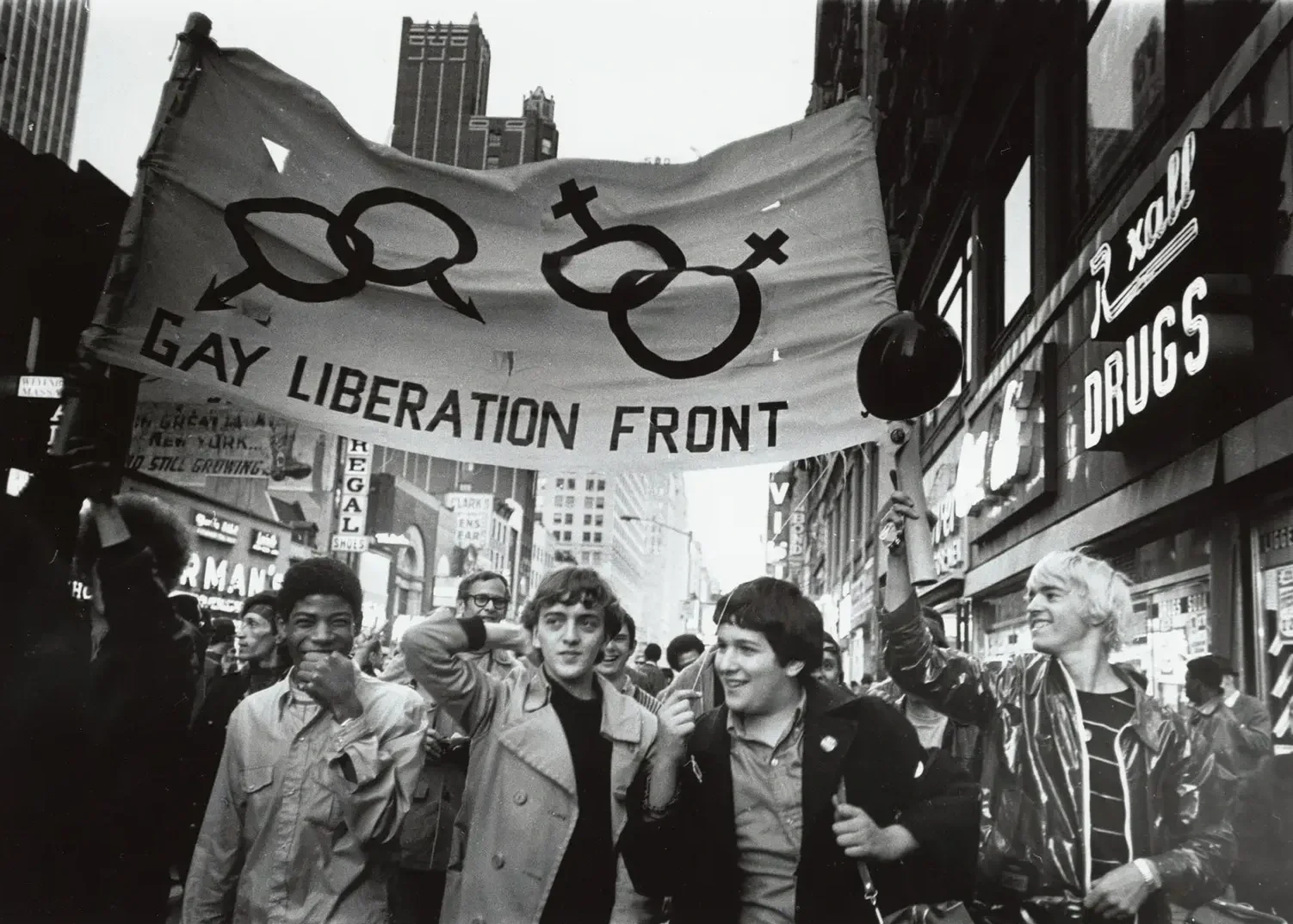 A black-and-white photo of a pride parade with a group of diverse young people walking in the street, holding a large banner that reads 'GAY LIBERATION FRONT' with symbols for different genders. The crowd is surrounded by tall buildings and storefronts, a person on the right with blond hair is smiling, and other participants are visible in the background.