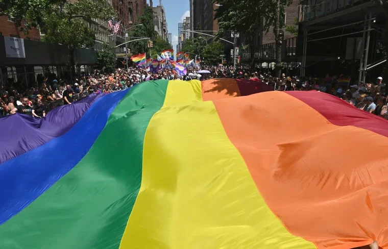 Large rainbow flag being held by crowd during a pride parade in an urban street, with many participants and onlookers, and additional rainbow flags and banners in the background.