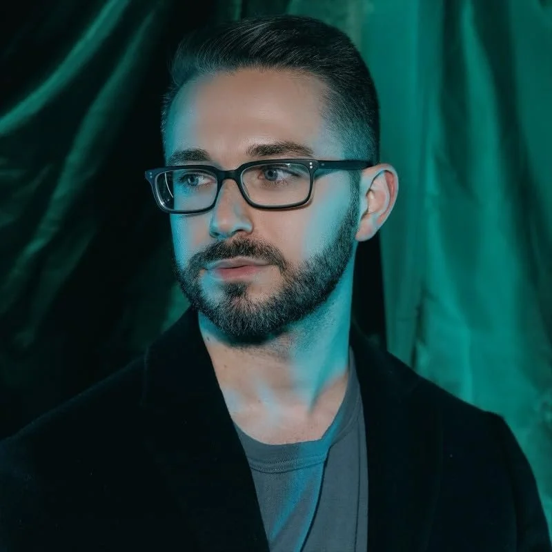A man with glasses and a beard looking to the side, with dark green drapery in the background.