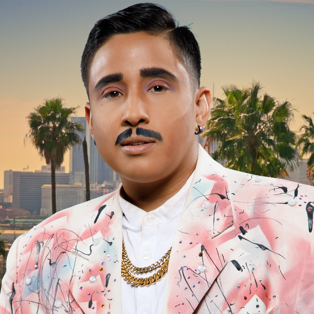 A person with dark hair, a mustache, and earrings wearing a pastel-colored jacket with abstract black, pink, and white patterns, and gold chains, standing against a background of palm trees and city buildings during sunset.