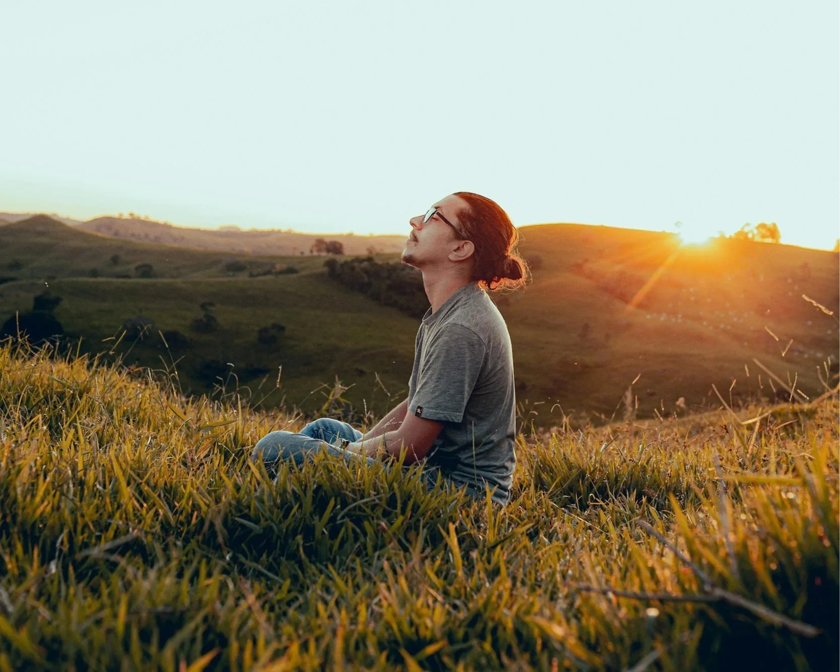 guy sitting a hilly field with his head towards the sky and eyes closed like he is at peace
