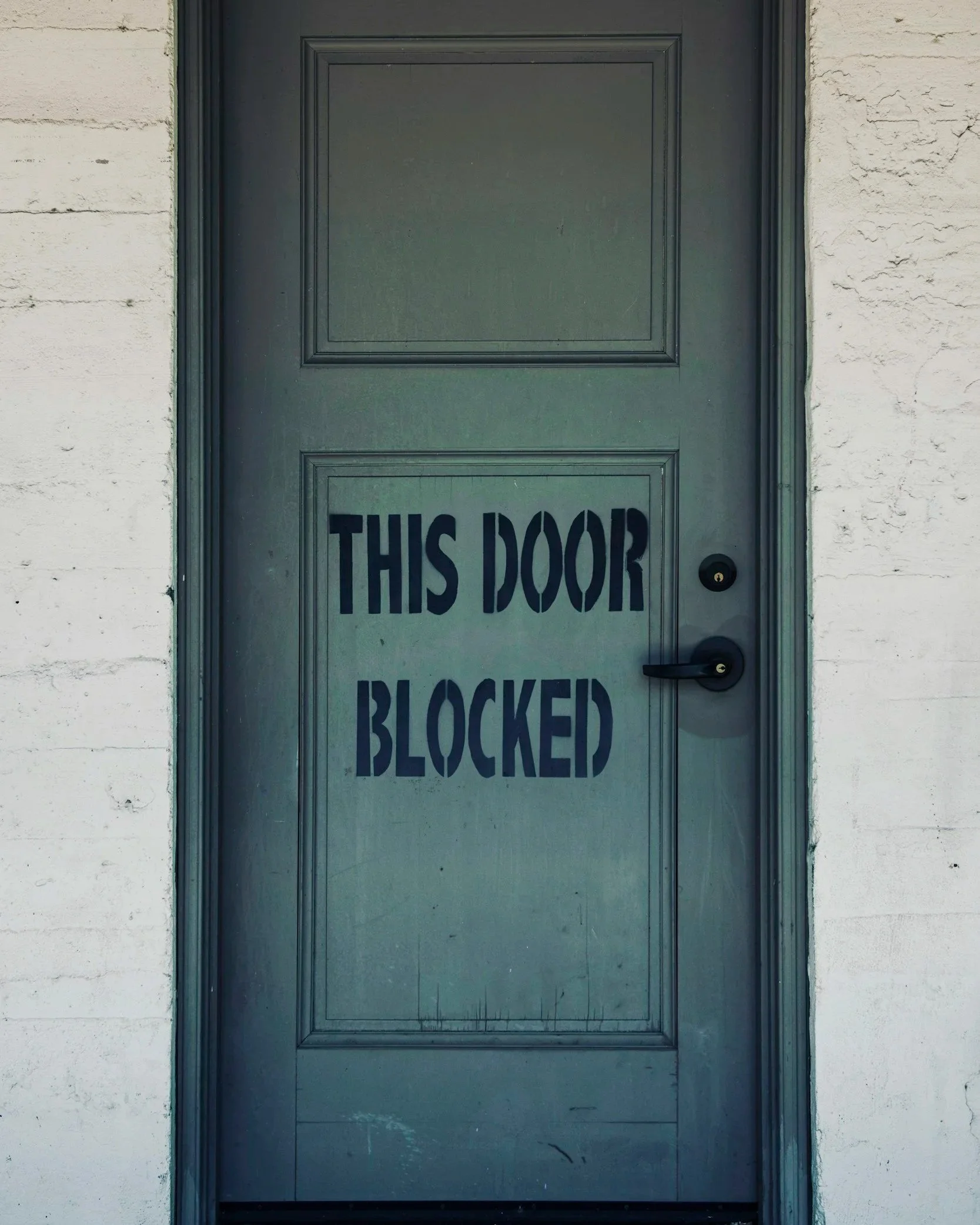 a green door that is shut with "this door blocked" painted on it
