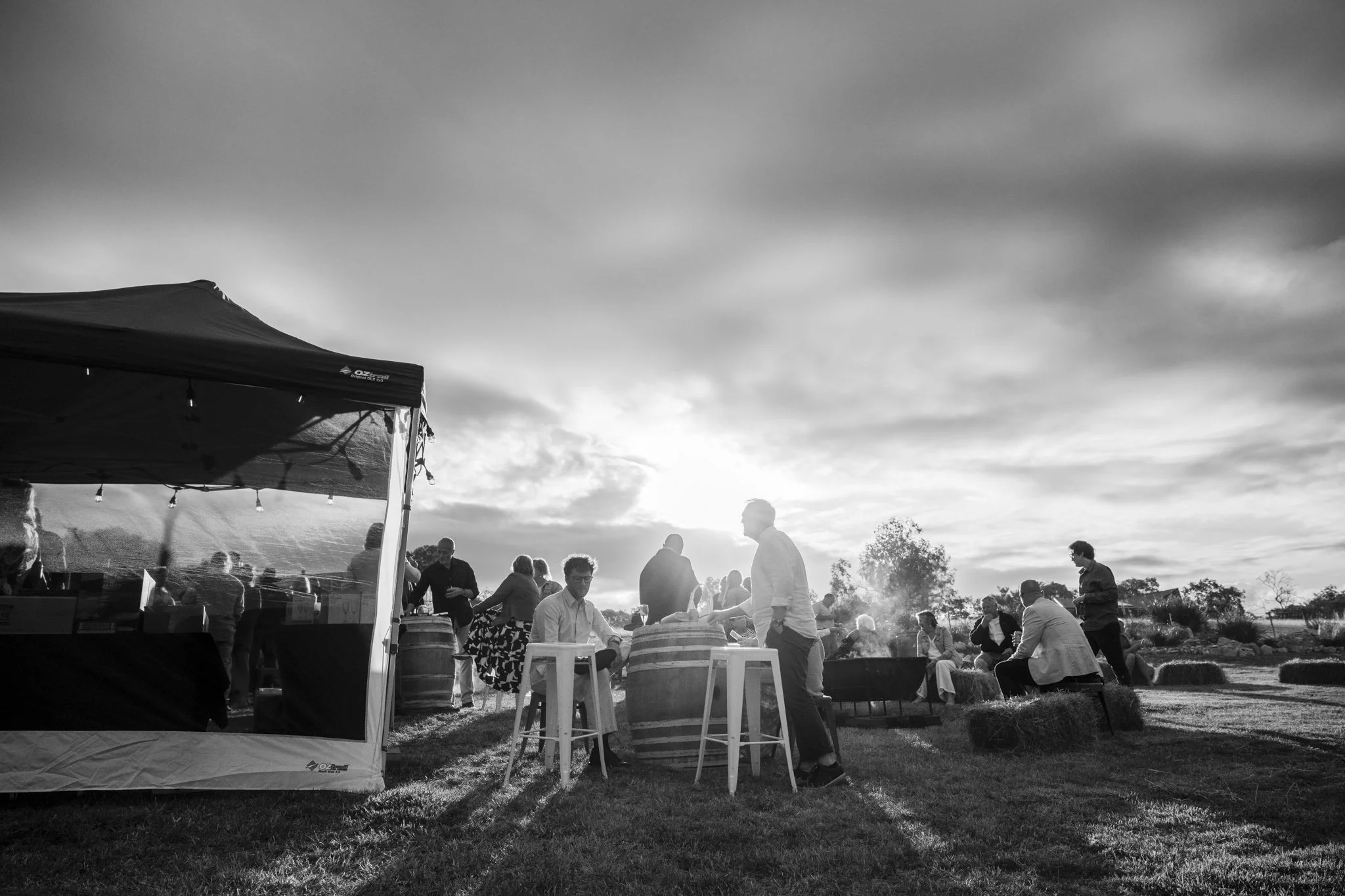 People gathered outdoors at a social event, some sitting on hay bales, others standing, with a tent on the left, in a rural setting during sunset with a cloudy sky.