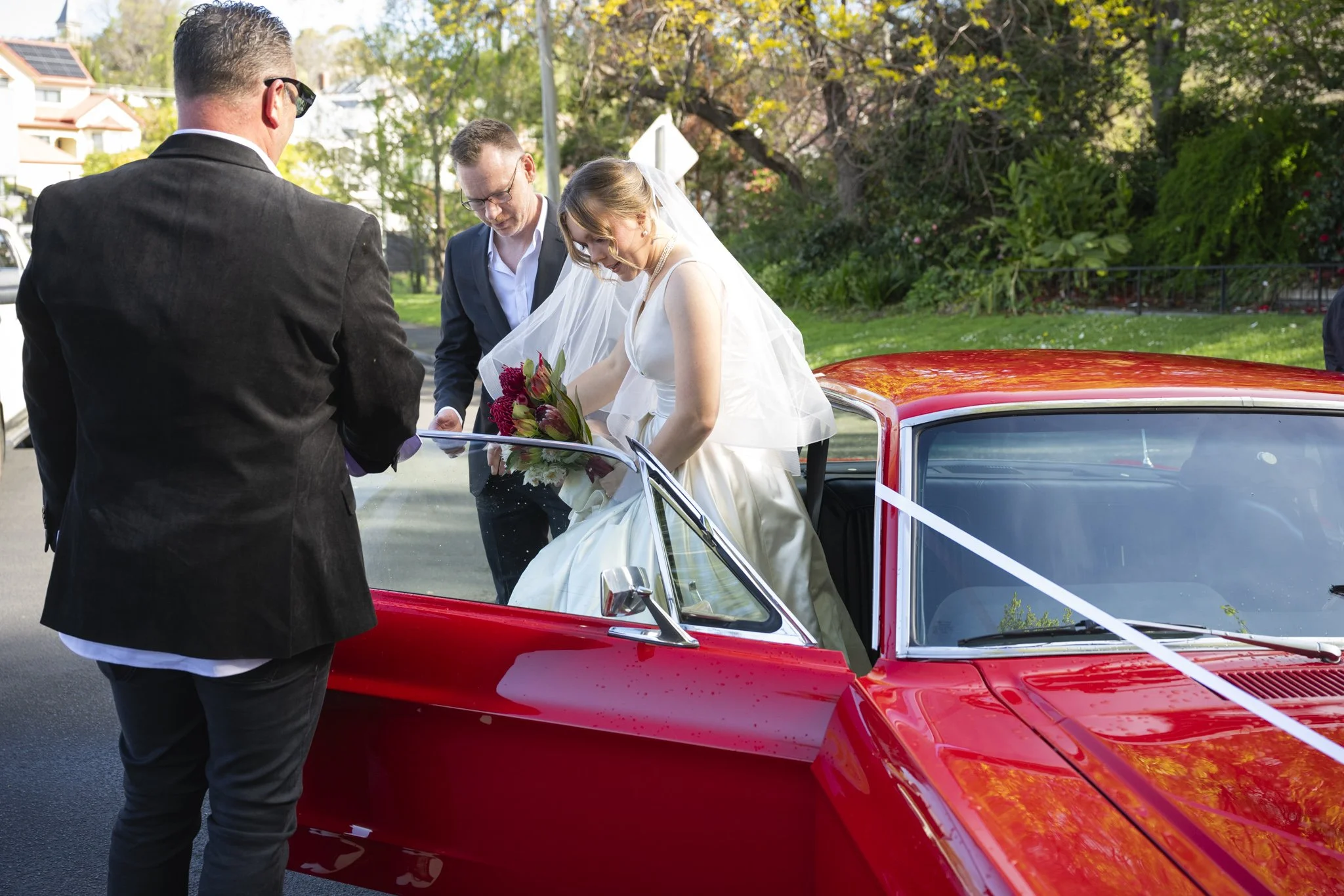 A bride in a white wedding dress getting into a red vintage car with her bouquet, as three men in formal suits look on outdoors during daytime.