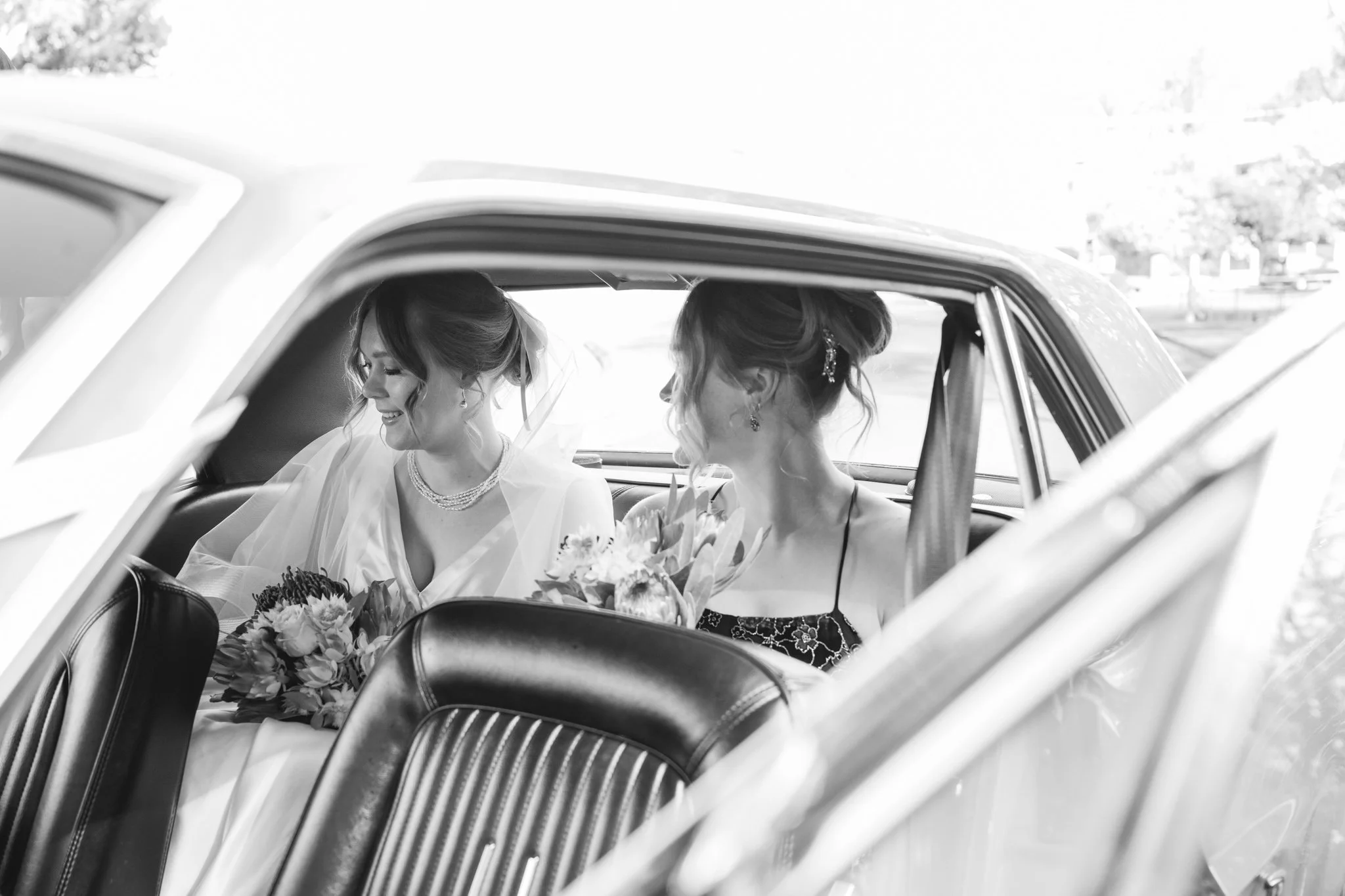 Two women sitting in the backseat of a classic car, dressed in wedding attire, smiling and holding bouquets of flowers.
