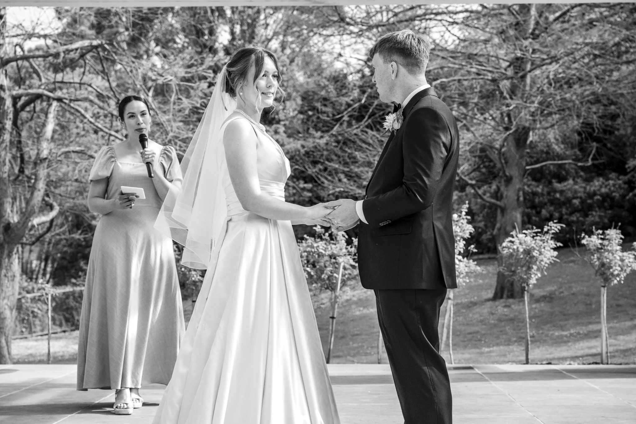 A black and white photo of a wedding ceremony outdoors, featuring a bride and groom holding hands and facing each other, with an officiant standing behind them holding a microphone.
