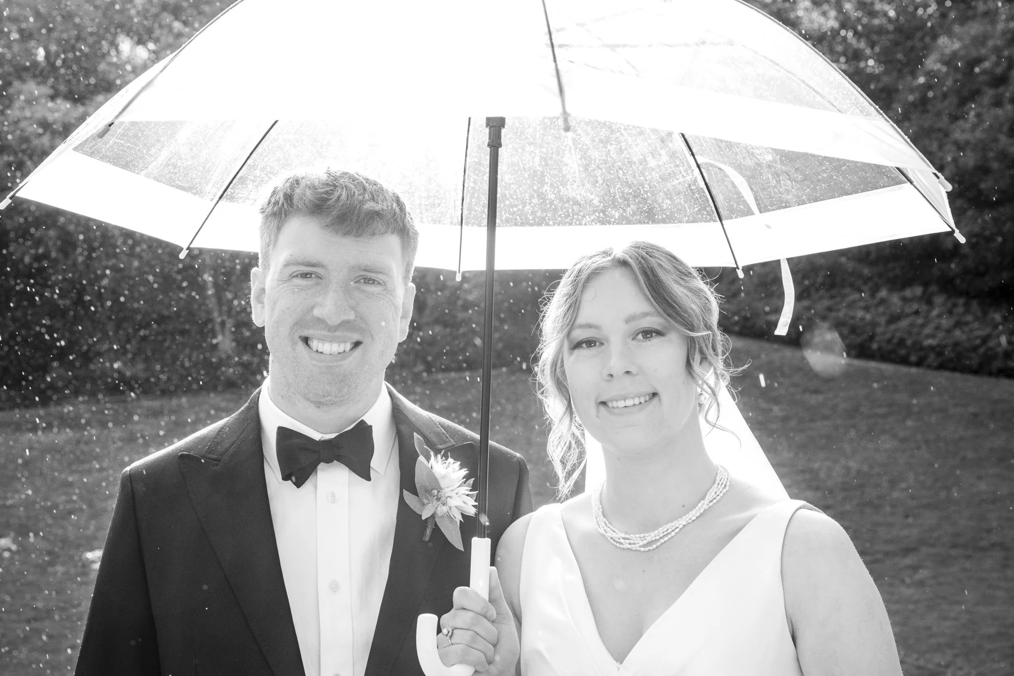 Black and white photo of a smiling bride and groom standing under a clear umbrella outdoors, with water droplets falling in the background.