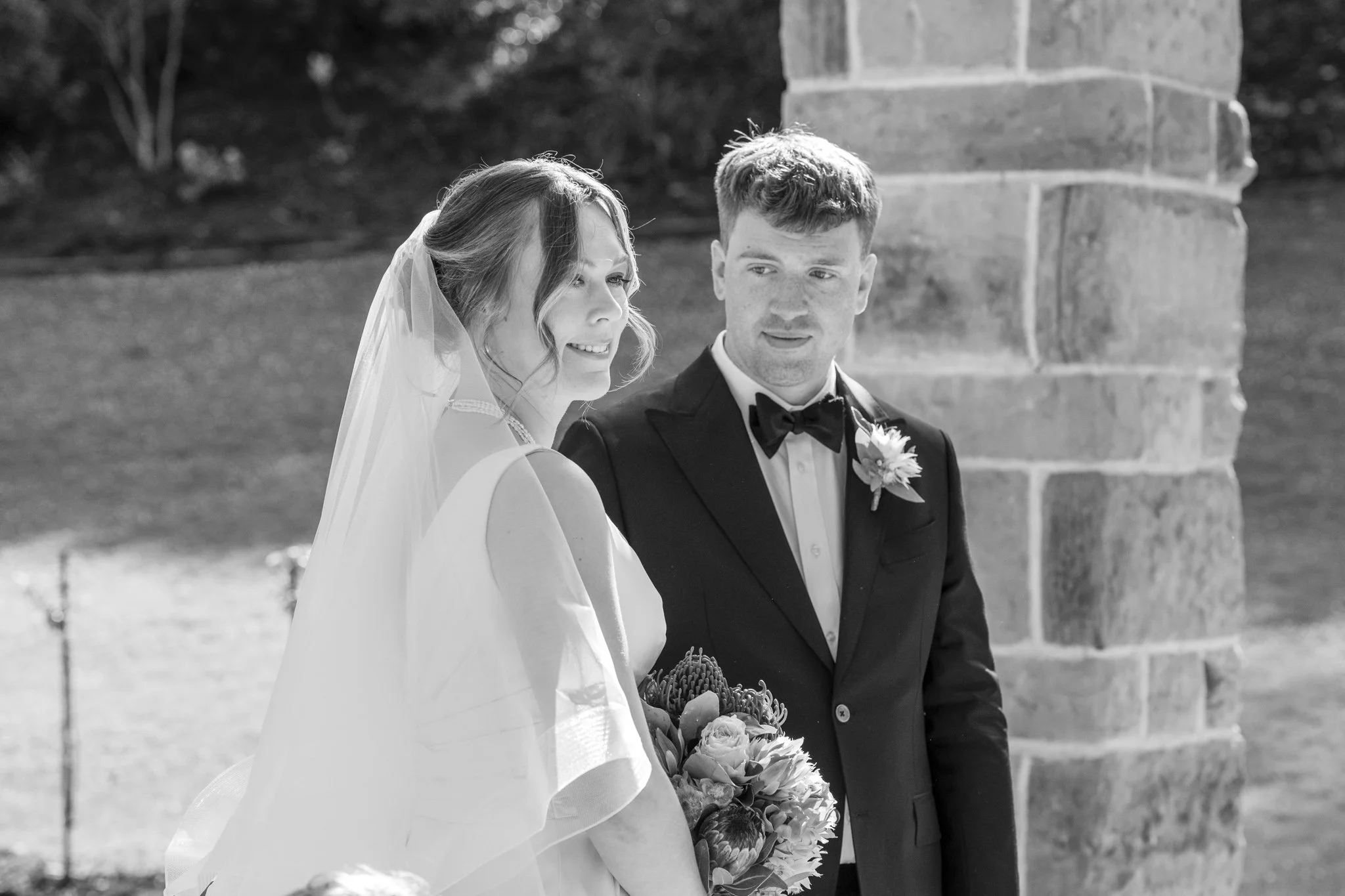 A bride and groom standing outdoors, with the bride holding a bouquet of flowers, during a wedding ceremony.