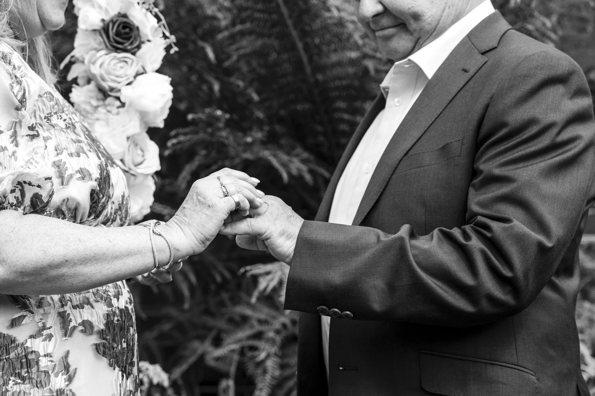 A man in a suit holding a woman's hand during a wedding or engagement ceremony outdoors, with a background of plants and flowers.