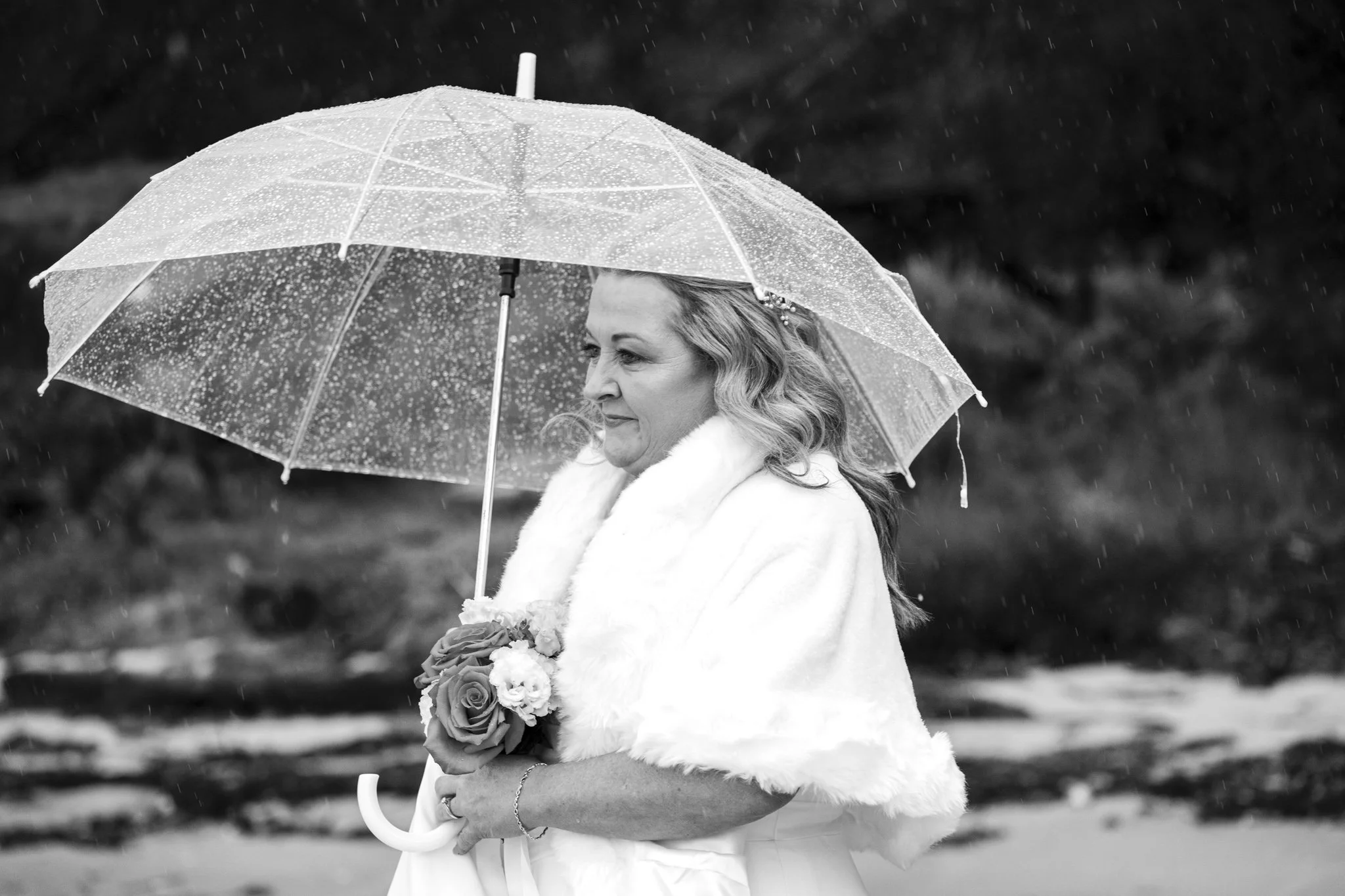 A woman in a wedding dress with a fur shawl holding a bouquet of flowers under a transparent umbrella during rain.