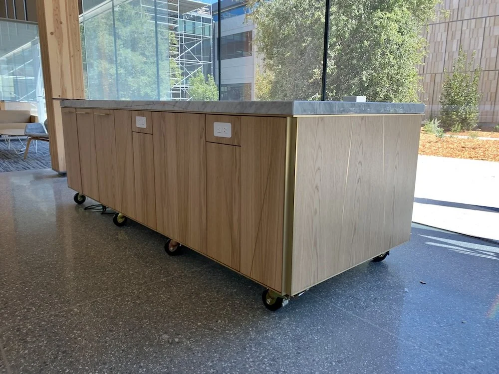 Custom mobile hospitality island with power on both sides, trash/recycle/compost disposal, deep storage cabinets for hospitality equipment, and brass inlay detailing.