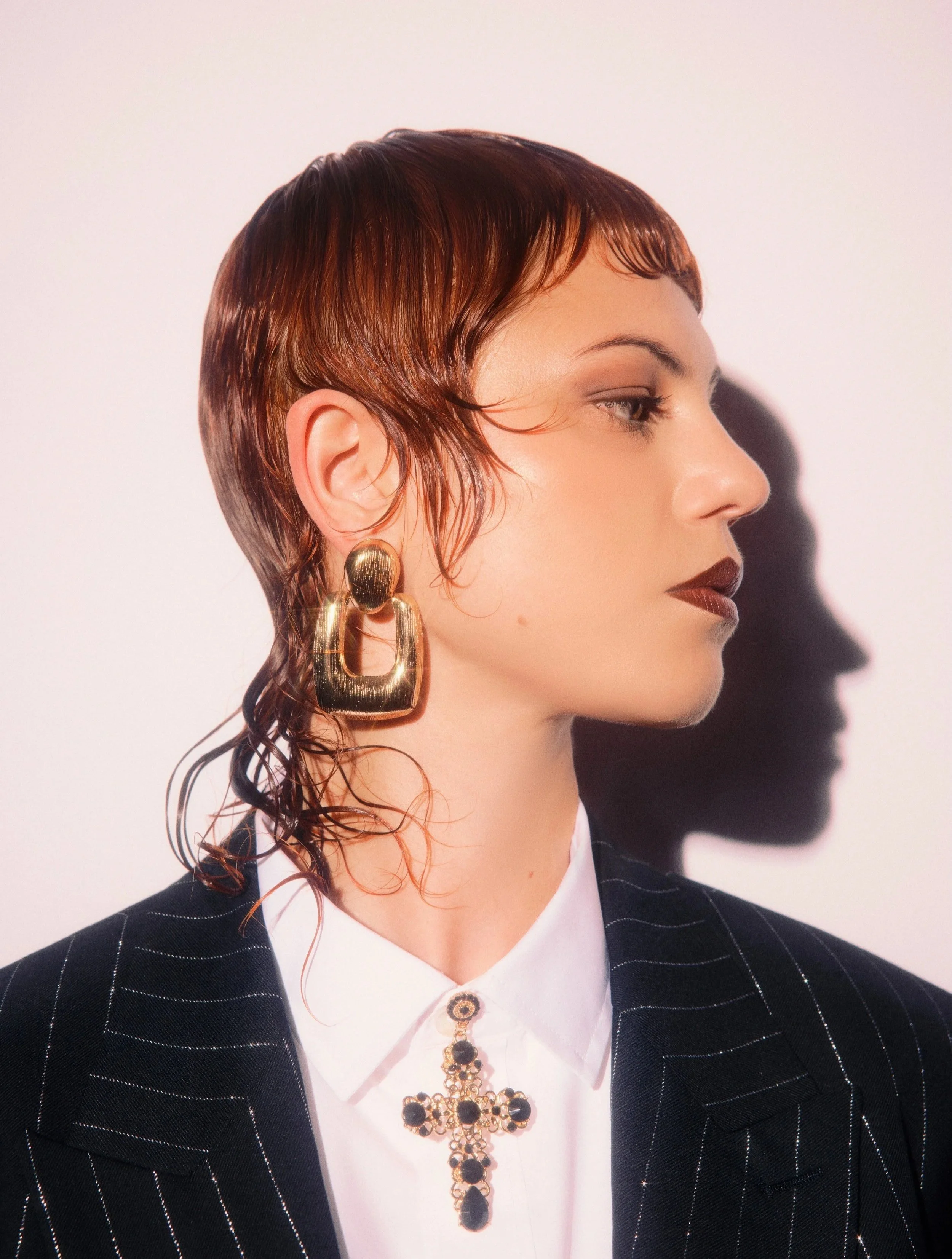 Side profile of a woman with short, wet reddish-brown hair styled with waves, wearing large gold earrings, and a black and silver pinstripe suit over a white blouse with a decorative cross necklace.