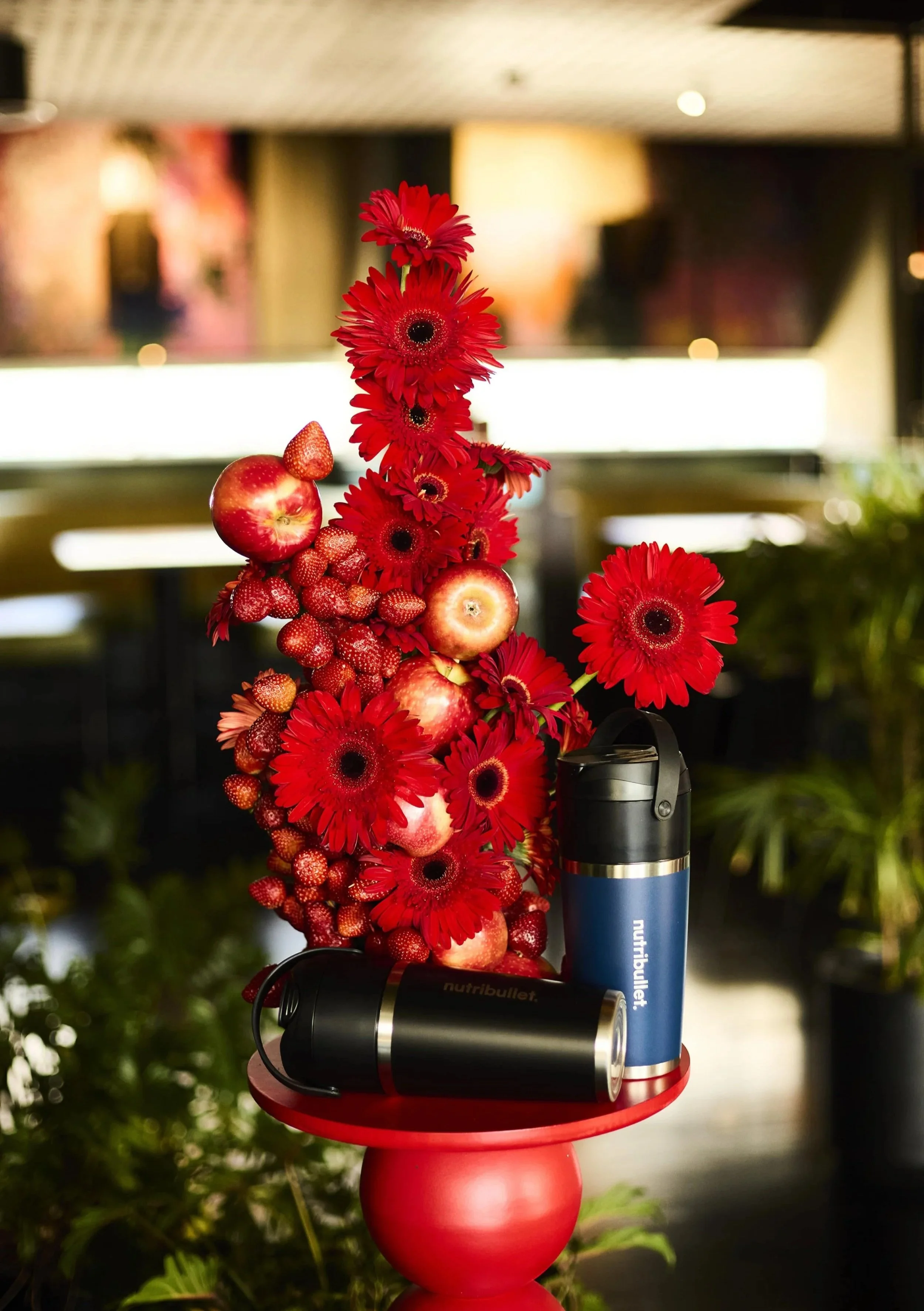 A festive arrangement with red gerbera daisies, strawberries, and apples on a red round table, with two black and blue NutriBullet blenders in front, set against a blurred indoor background.