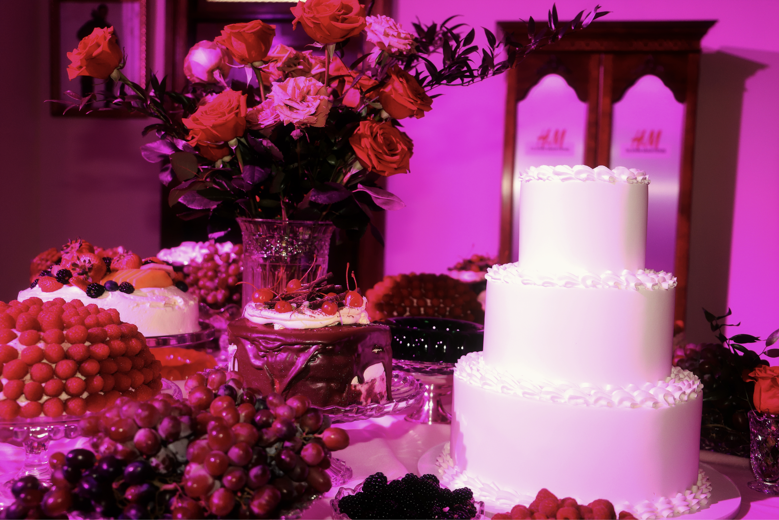 A dessert station, including a three-tiered white cake, decorated with white frosting, alongside various smaller cakes topped with berries and fruit, and a large bouquet of pink and red roses in a glass vase, all under pink lighting.