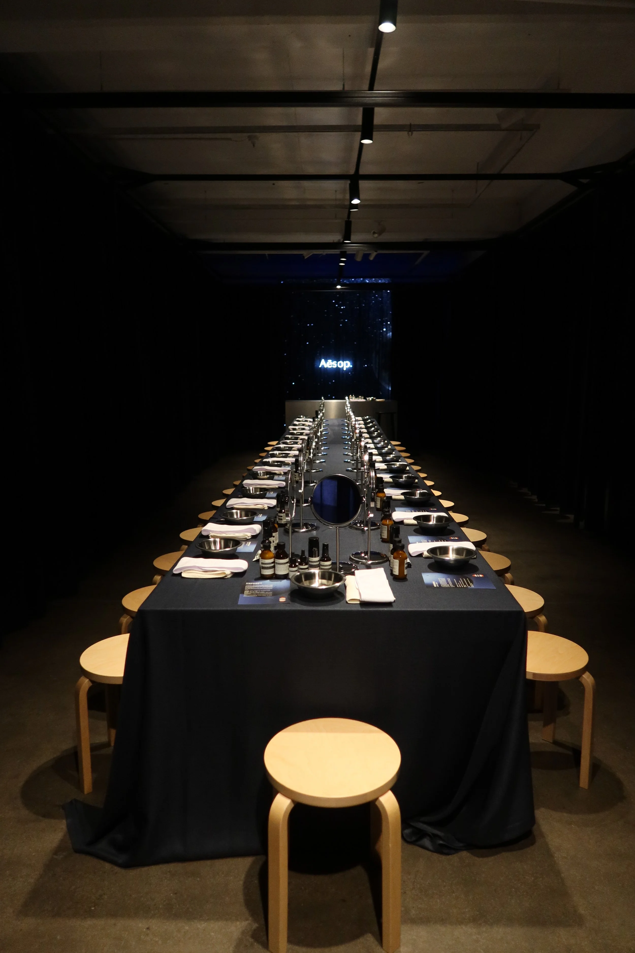 A long table set for a skin care workshop with navy tablecloths, small bowls, chrome mirrors, and bottles, flanked by small wooden stools in a dimly lit room with navy draped walls, and an LED screen at the end displaying the word 'Aēsop'.