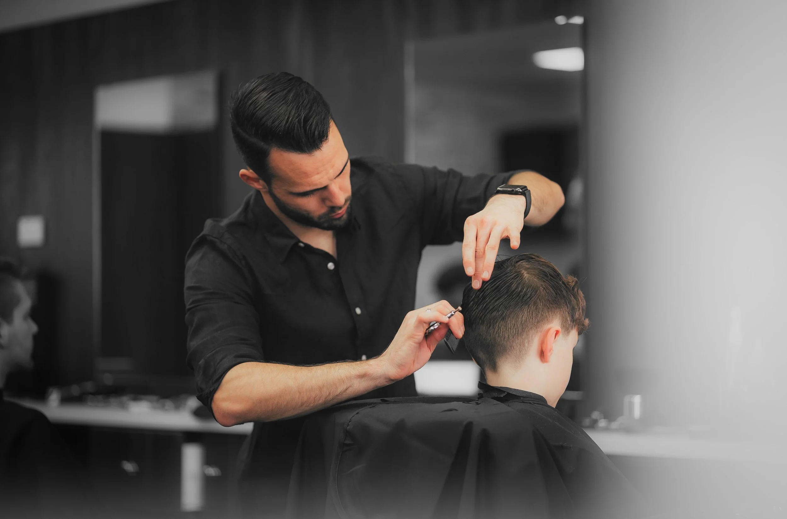 A barber is cutting a young man's hair in a salon. The barber is concentrating while working with scissors and a comb, wearing a black shirt. The young man is seated, facing away from the camera, with a haircut in progress.