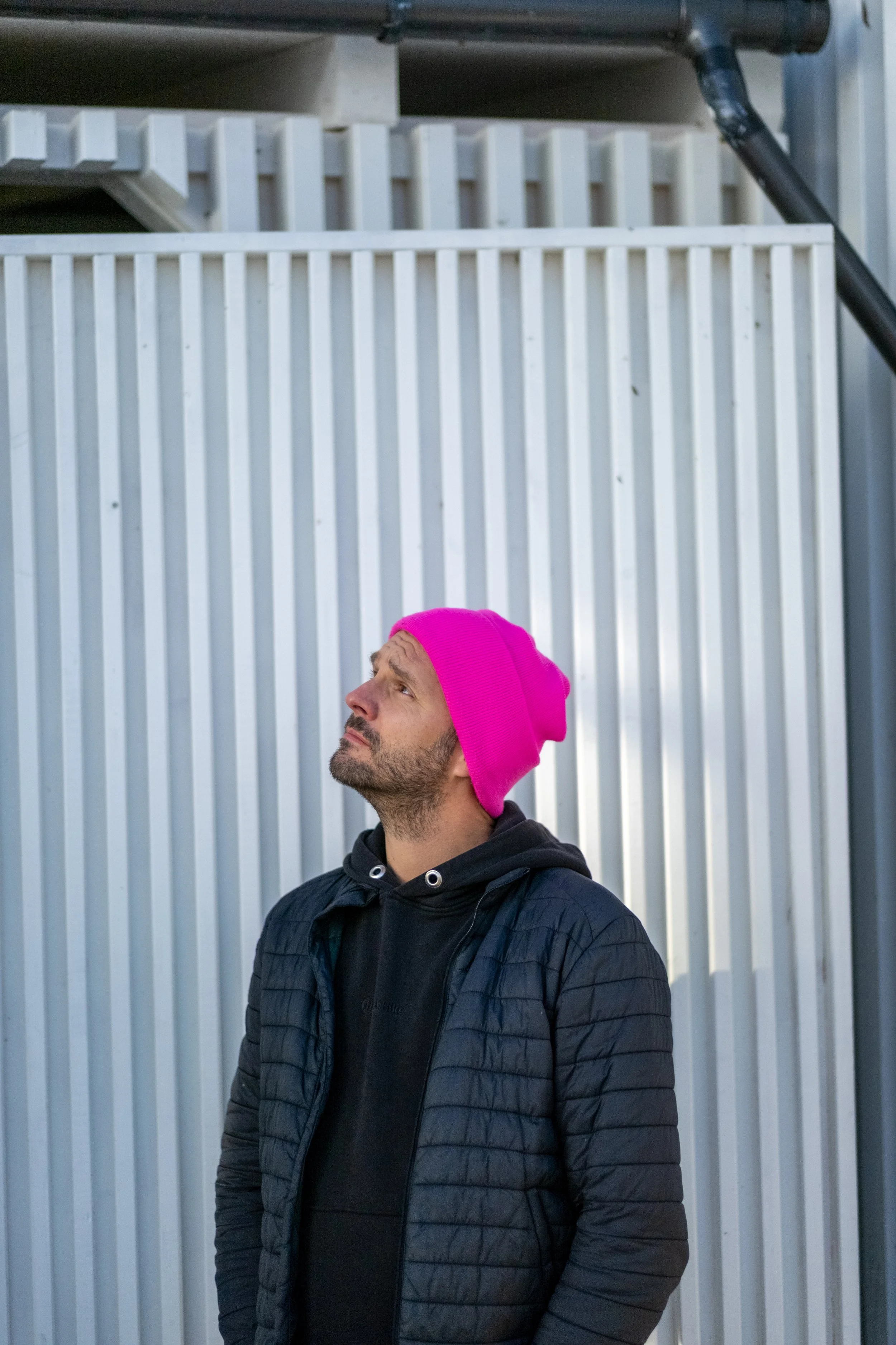 A man wearing a bright pink beanie and a black jacket looks up at a pipe or vent above him against a metal wall.