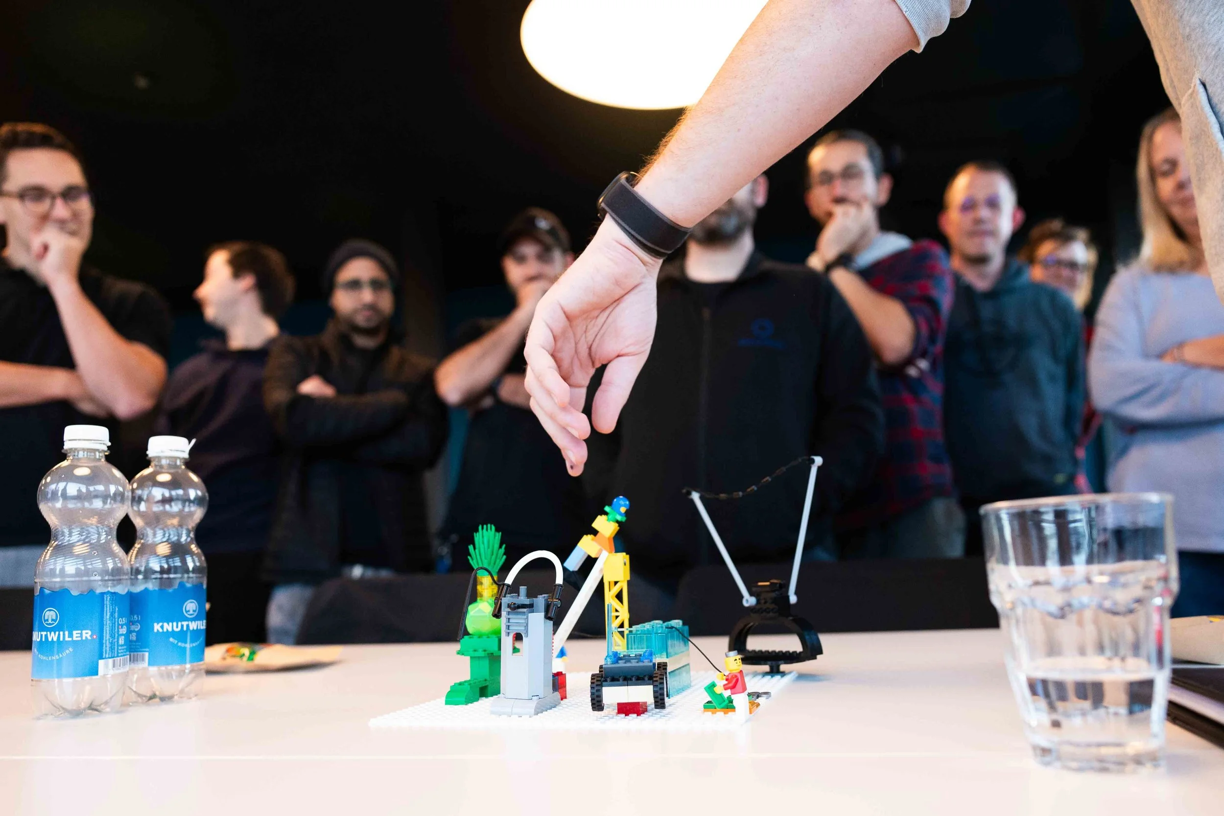 Person demonstrating a LEGO Rube Goldberg machine to an engaged audience in a dark room with a large round light overhead.