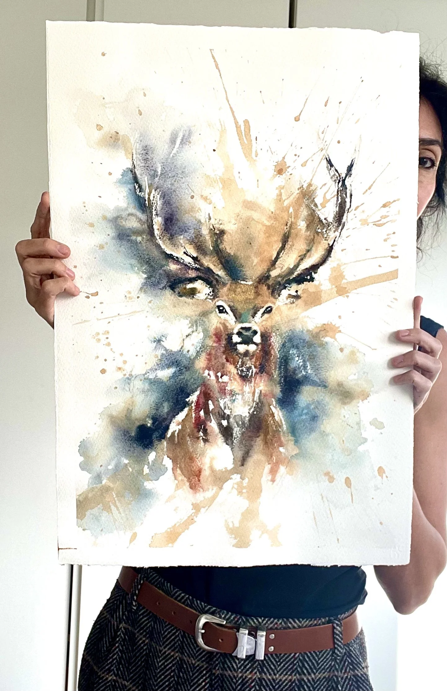 Person holding a watercolor painting of a deer with large antlers, rendered in vibrant and splattered paint style.