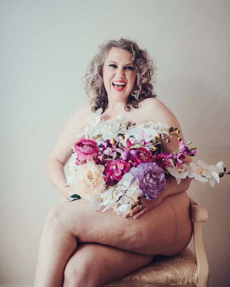 A woman with curly gray hair smiling and holding a large bouquet of various colorful flowers, sitting on a chair with her legs crossed.
