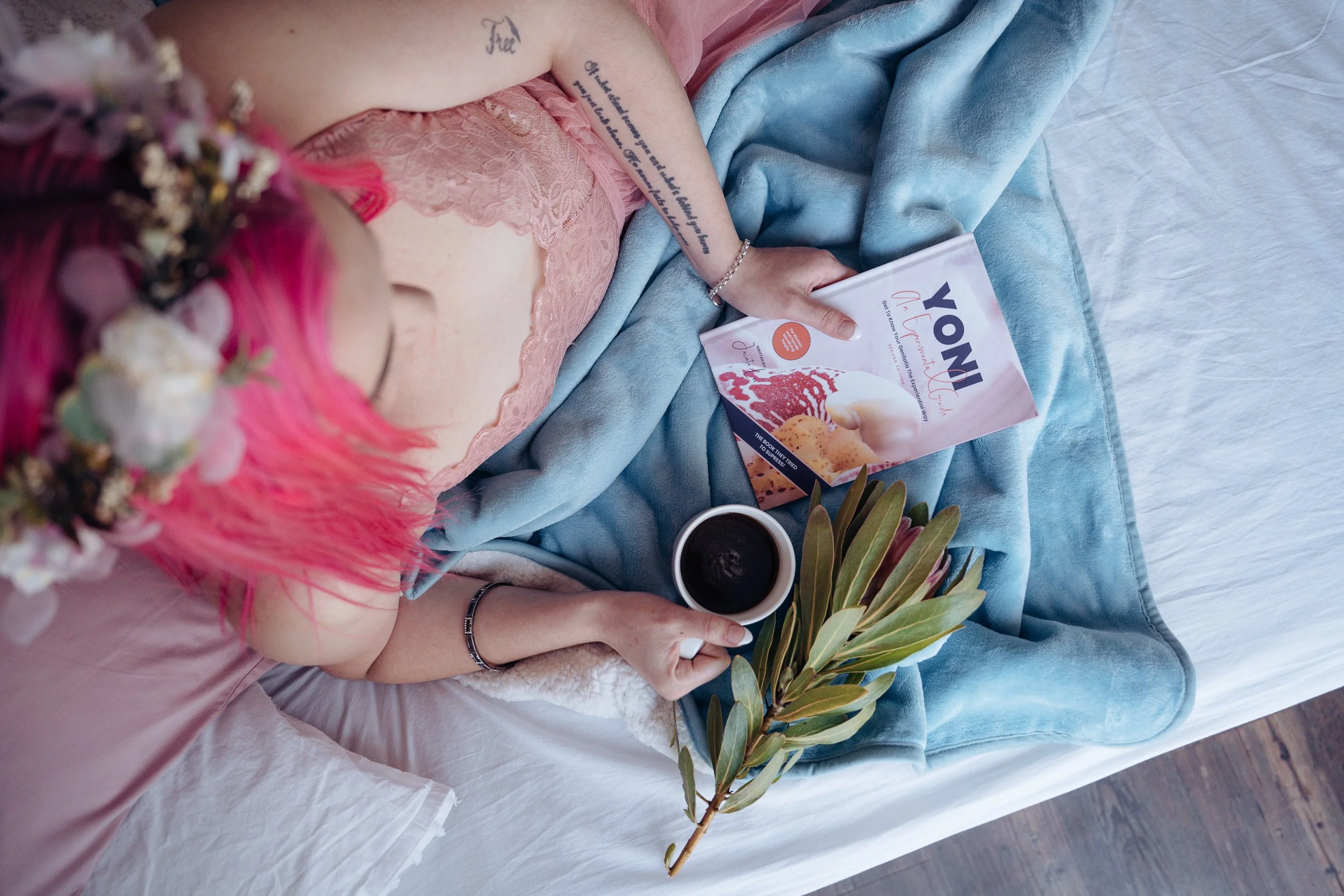 A woman with pink hair and a flower crown lying on a bed, holding a cup of coffee in one hand and a magazine in the other. There is a blue blanket, a book titled 'VONI', and some green leaves on the bed.