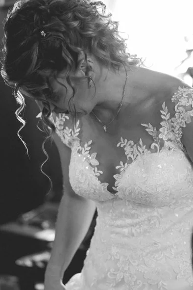 A woman in a wedding dress with floral lace detailing on the shoulders and bodice, looking downward with curly hair styled in loose waves.