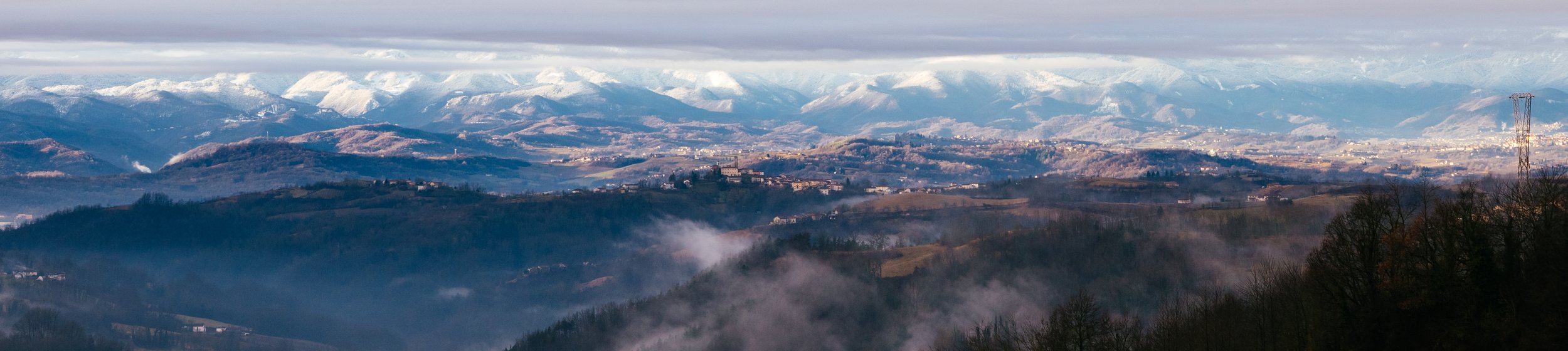 Landscape of mountain range with snow-capped peaks, rolling hills, forests, and low clouds or fog.