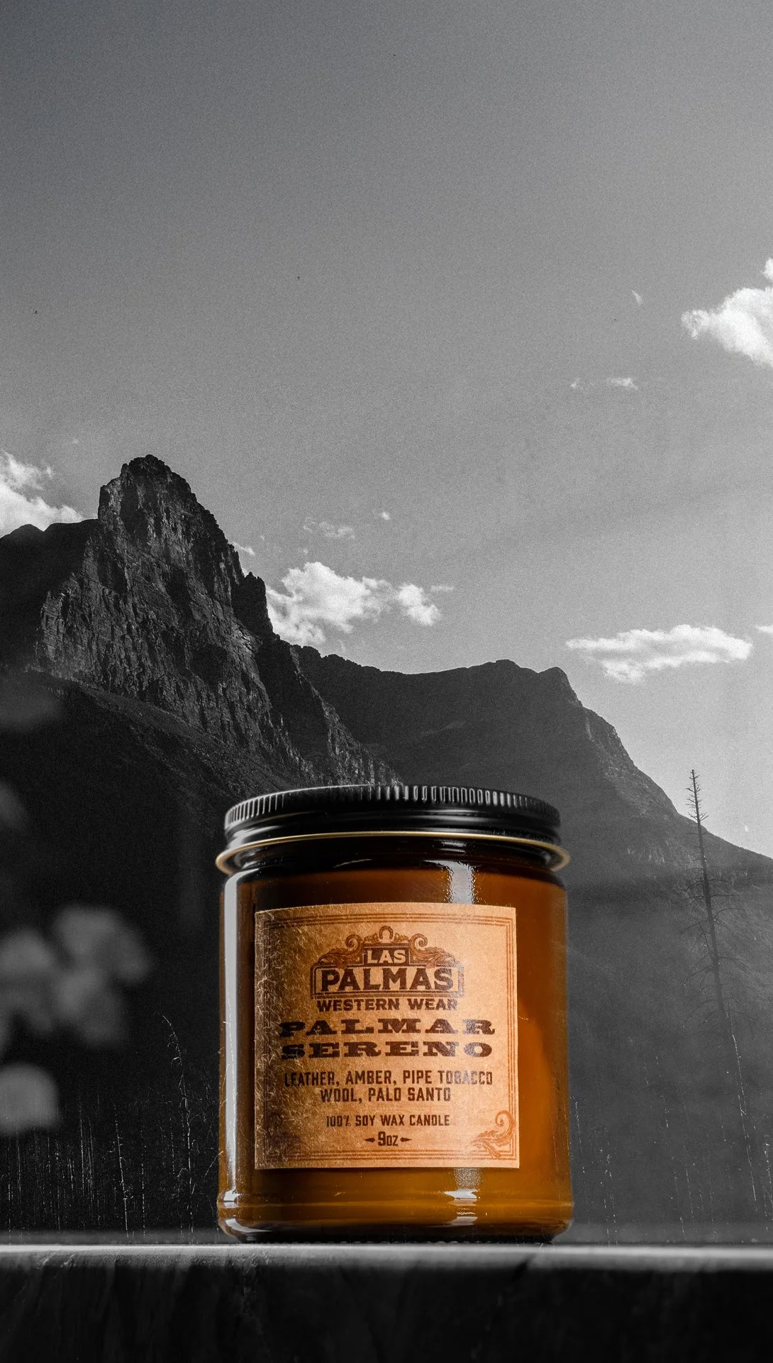 A brown scented candle with a label reading 'Las Palmas Western Wear Palmaerno' sitting outdoors in front of a mountainous landscape with a partly cloudy sky.