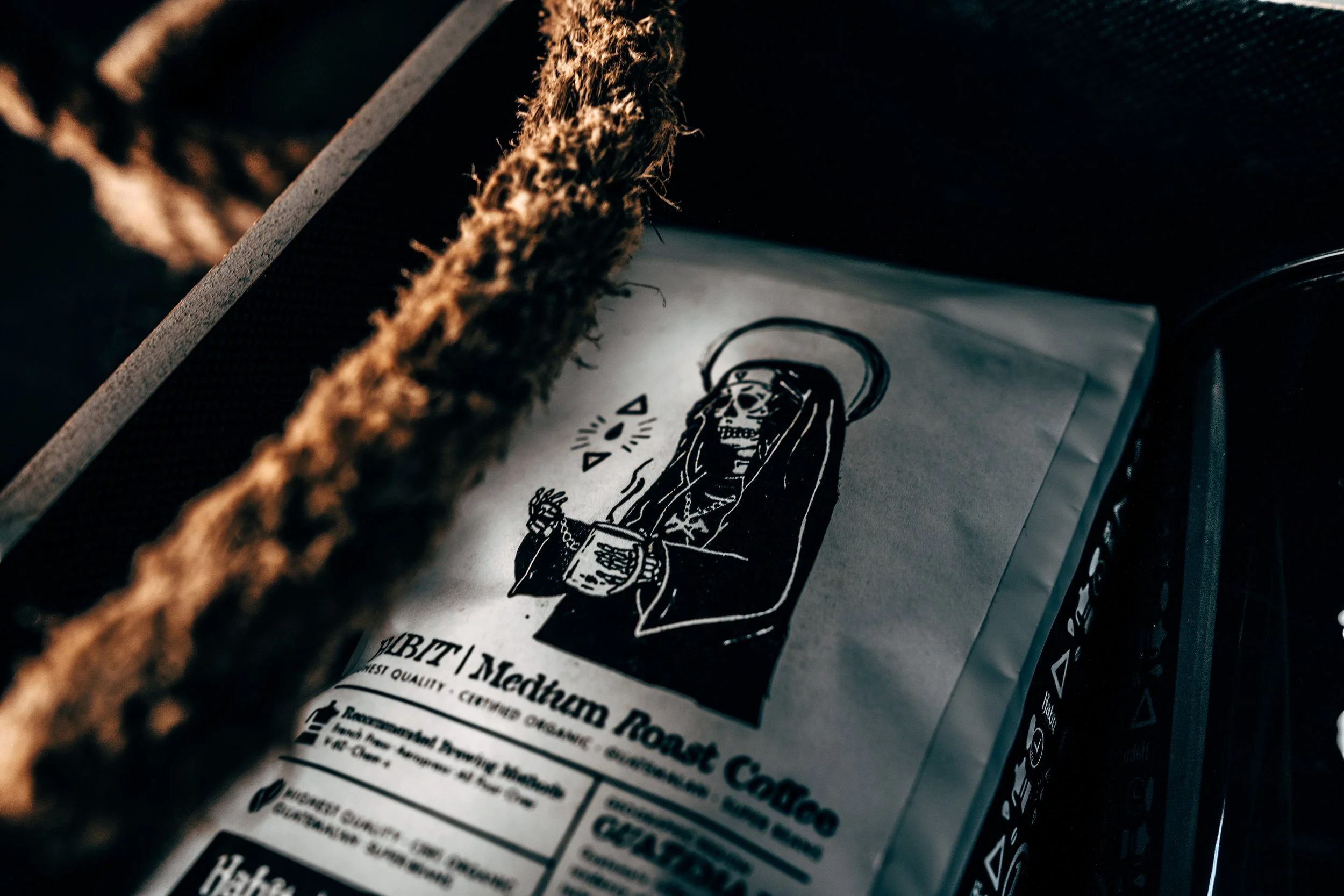 Black and white image of a bag of coffee with a drawing of a skeleton dressed as a saint, holding a cup. The text on the bag reads 'Subt | Medium Roast Coffee' and mentions quality and origin details.