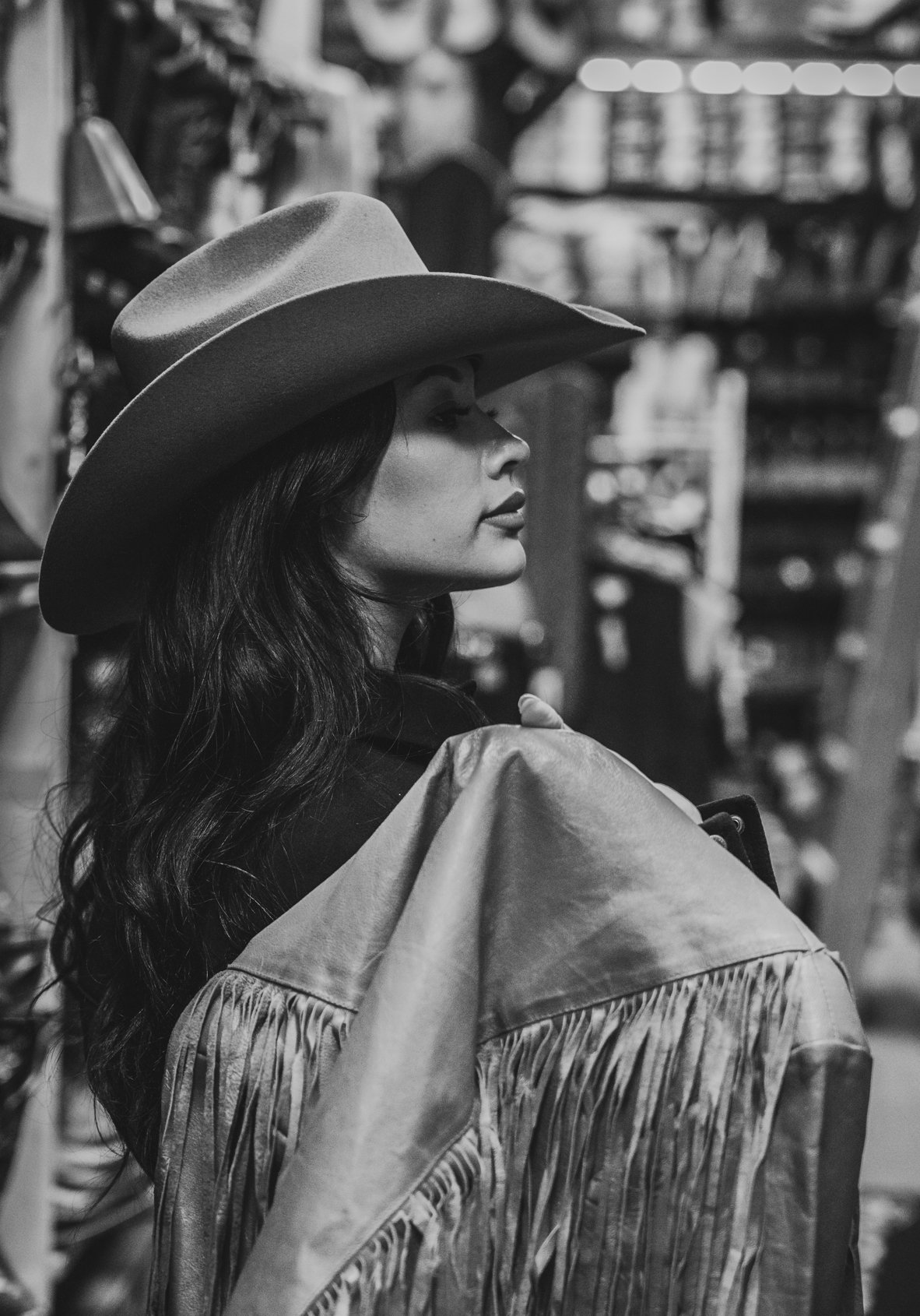 A woman with long dark hair wearing a wide-brimmed hat and a fringed jacket in a store or shop, captured in side profile in black and white.