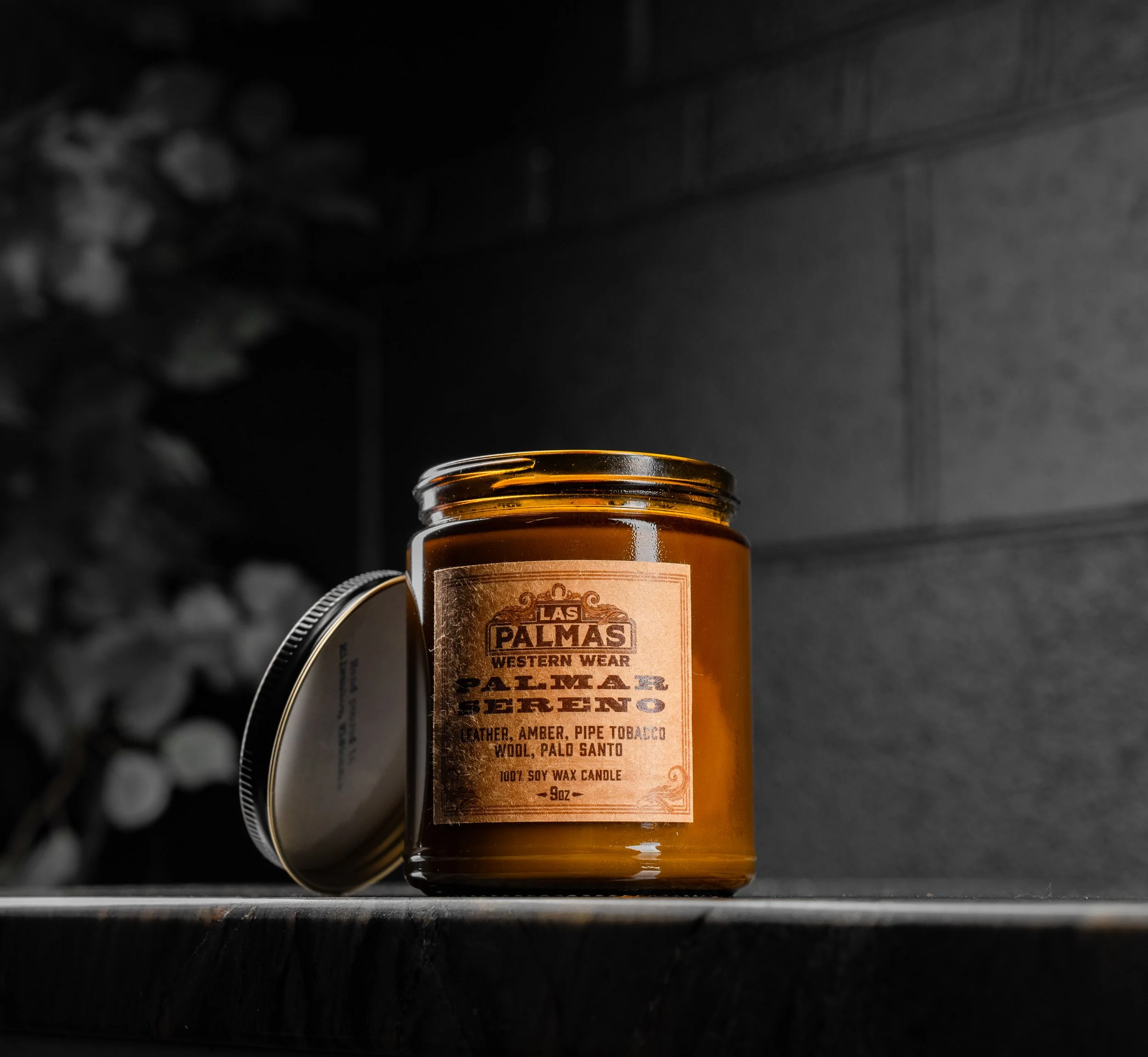 A brown candle jar with a label reading 'Las Palmas Western Wear Palmar Pepper, leather, amber, pipe tobacco, wool, Palo Santo, 100% soy wax, 9oz' sit on a dark surface with a slightly opened metal lid leaning against the jar, against a dark backgrou