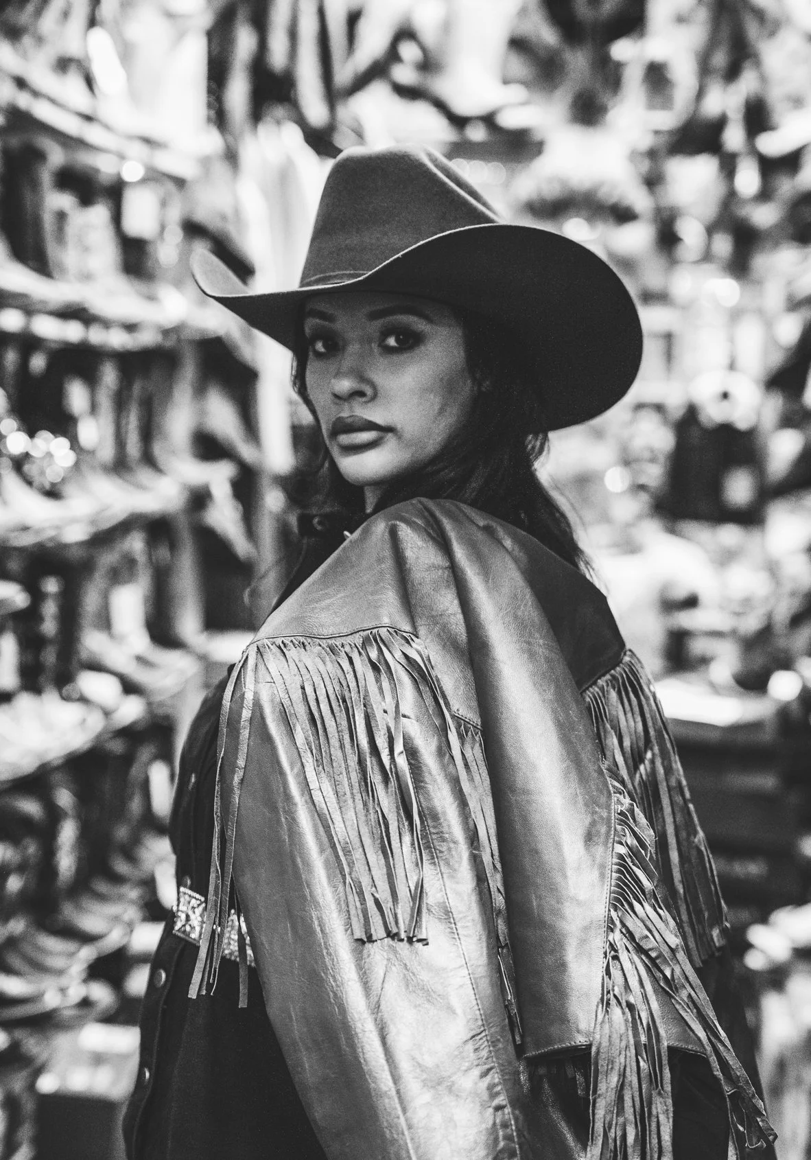 A woman wearing a cowboy hat and leather jacket with fringe, looking over her shoulder in a store with shoes on shelves in the background.