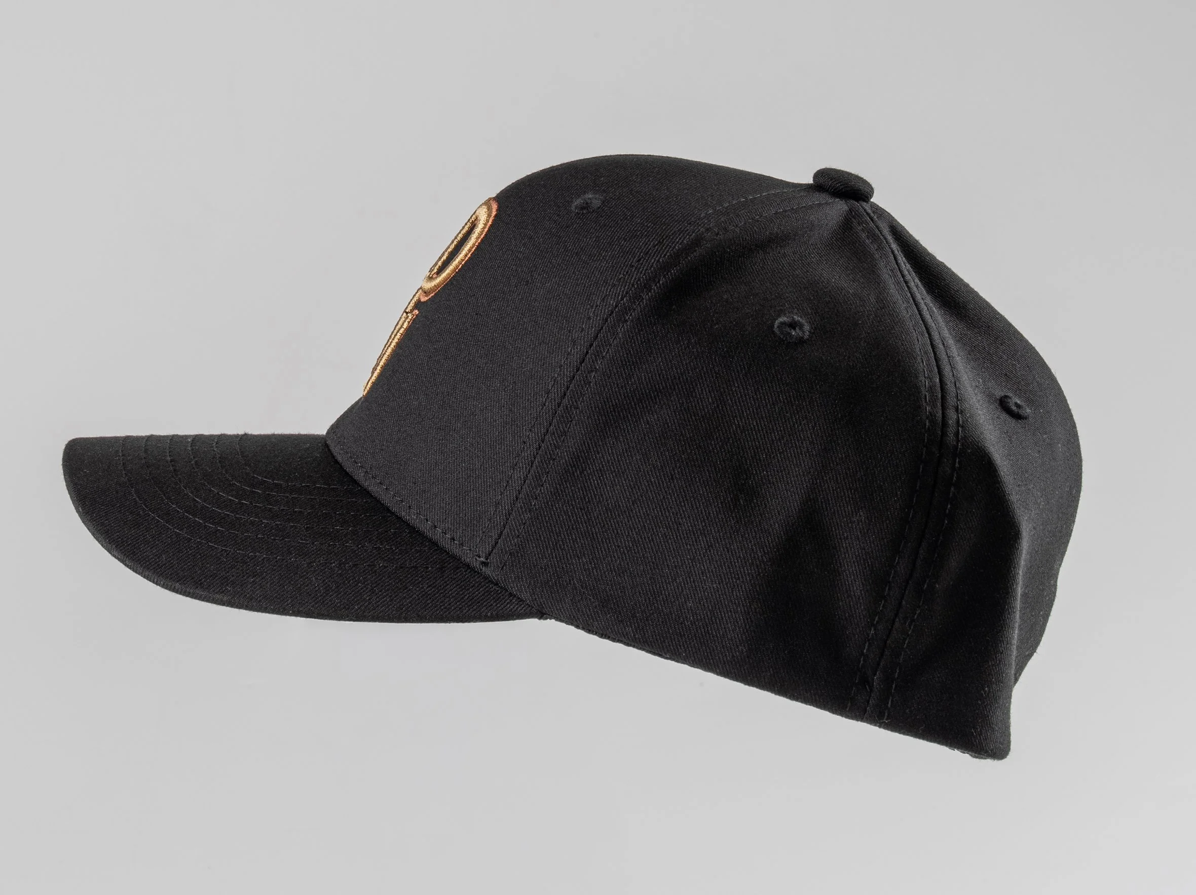 Black baseball cap with gold embroidered logo on front, facing sideways on light gray background.