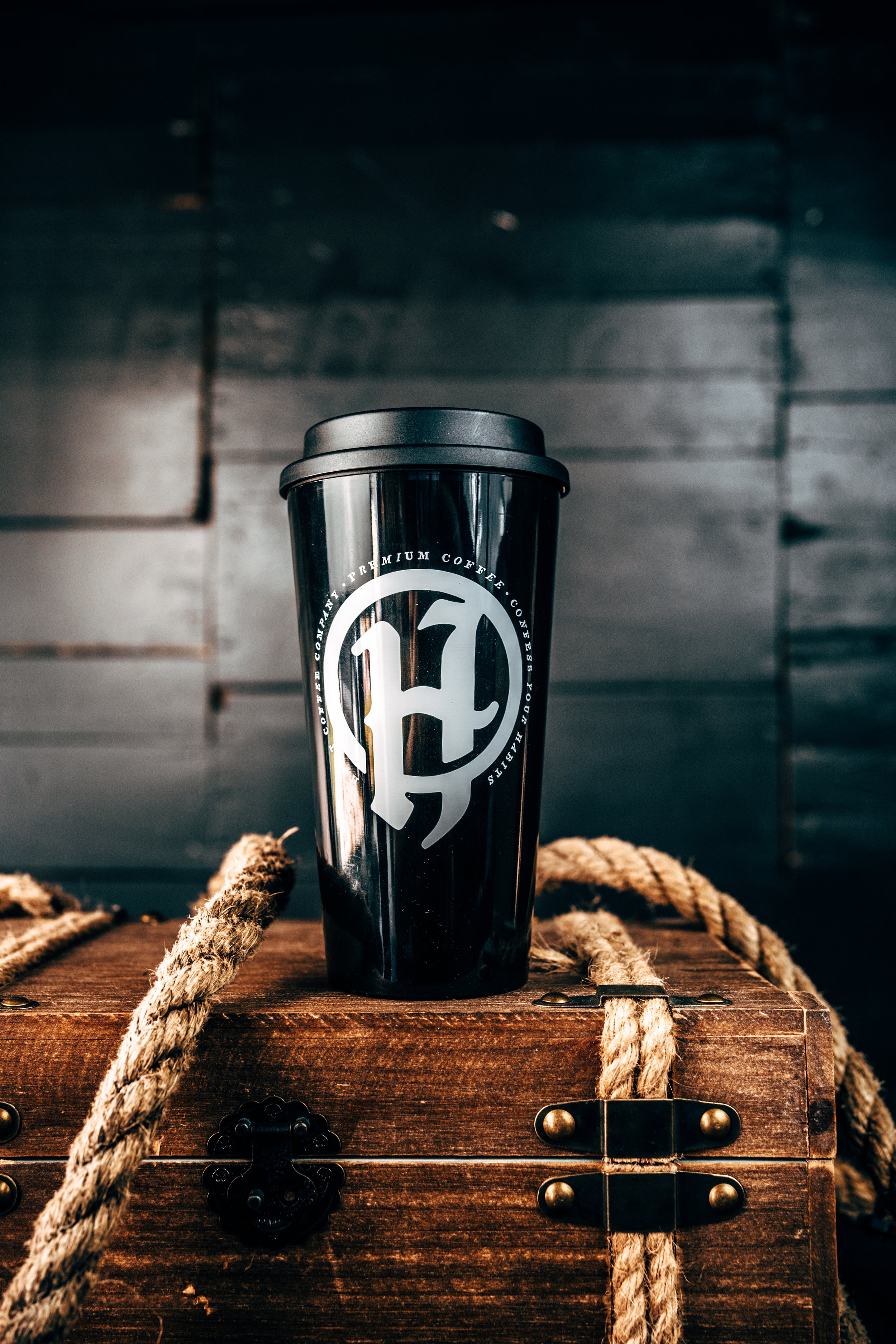 A black travel mug with a logo, sitting on a wooden chest with rope details, against a dark wooden background.