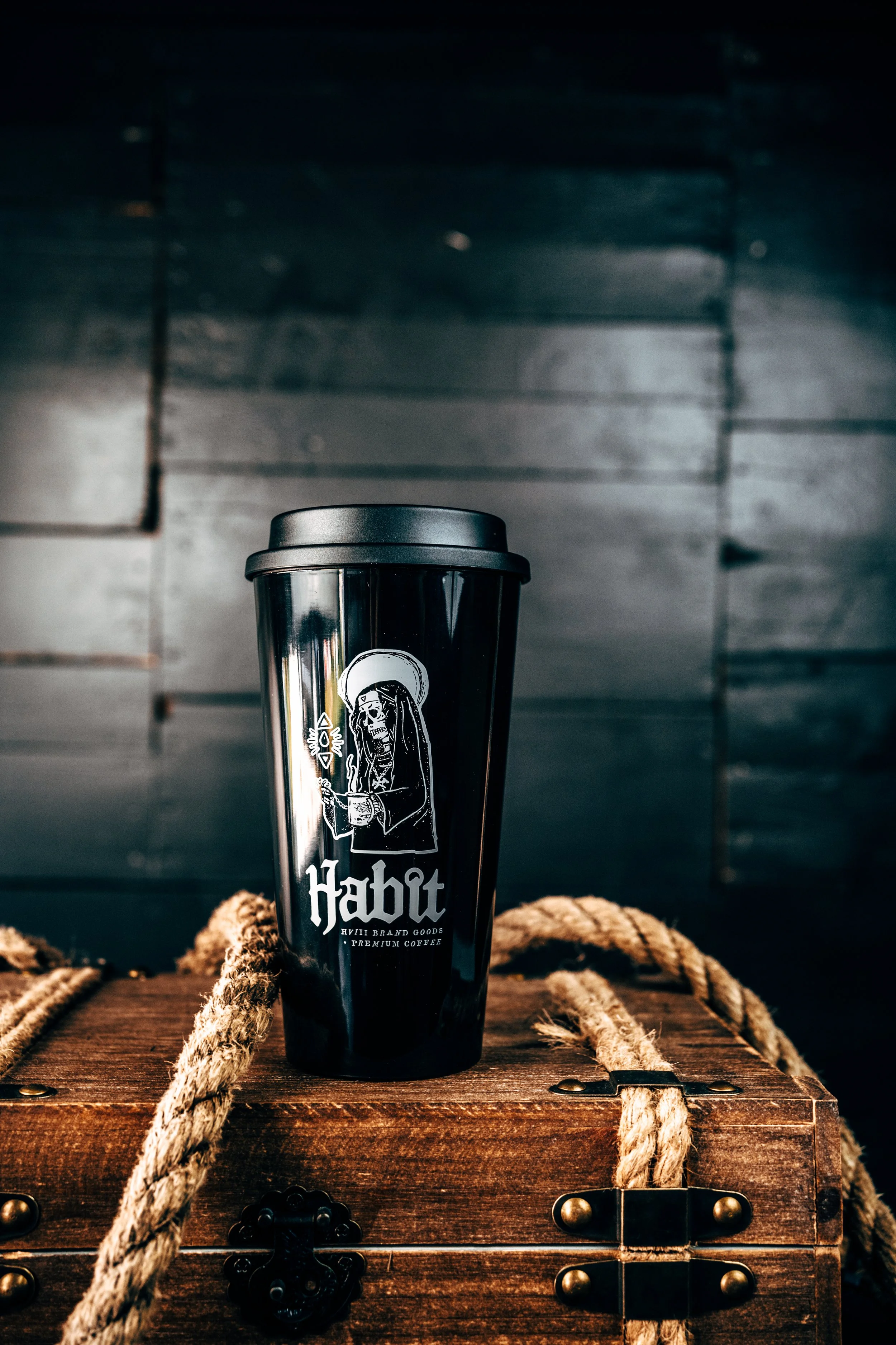A black reusable coffee cup with a skeleton holding a mug and a halo, labeled 'Habat' with smaller text 'Hyli Brand Goods Premium Coffee,' placed on a wooden box with ropes, against a dark wooden background.