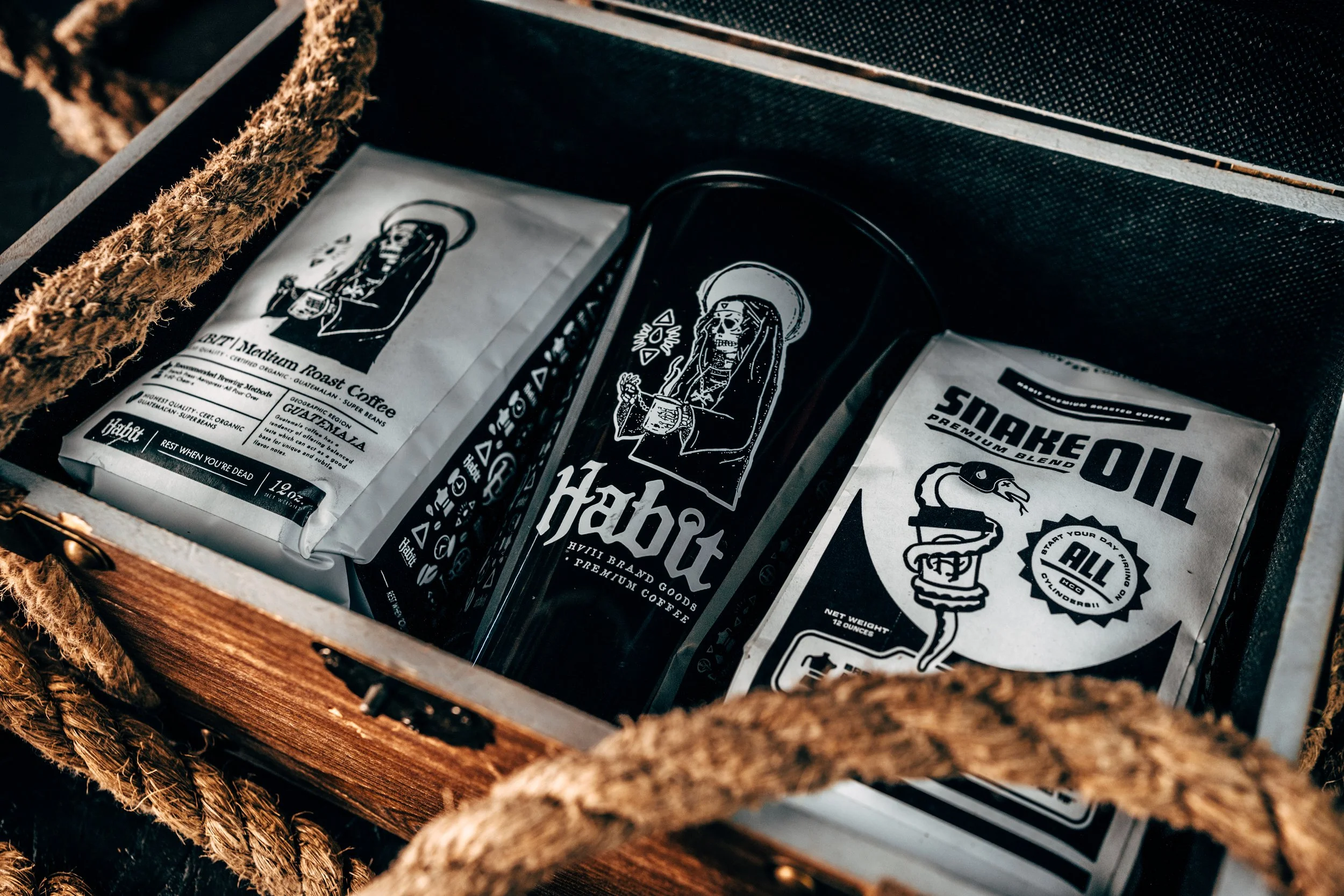 A wooden box containing three packages of coffee and a black travel mug. The packaging features black and white illustrations and text.