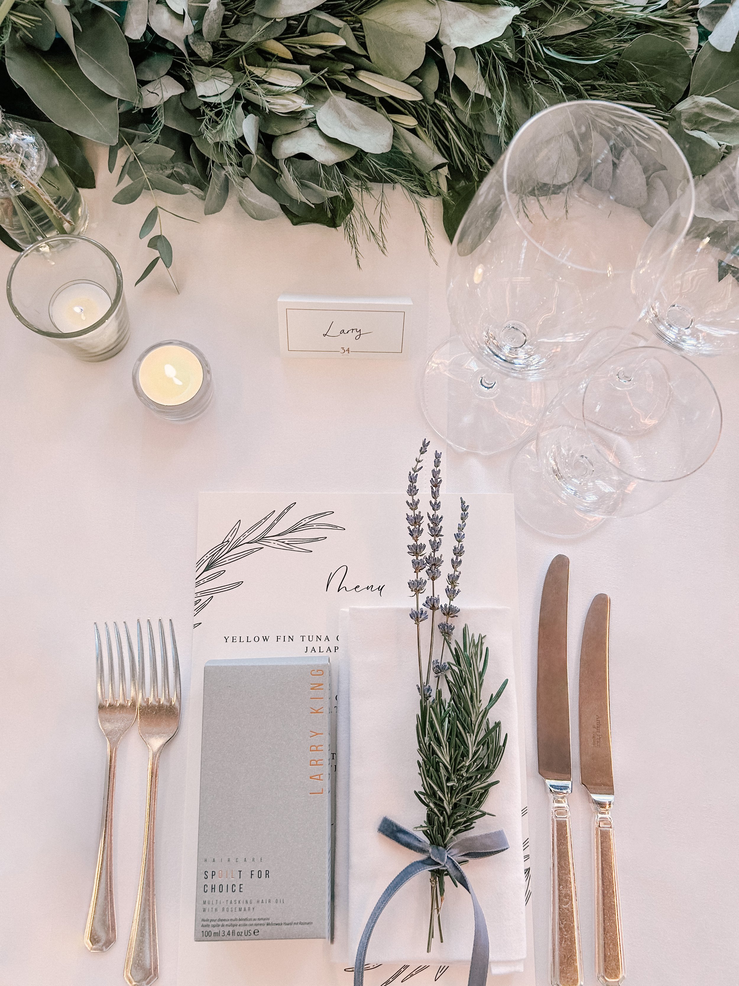 Elegant table setting with two forks, a butter knife and a butter spoon, lavender sprigs tied with a gray ribbon, candles, and a place card with the name Larry, number 34, against a backdrop of greenery and empty wine glasses.
