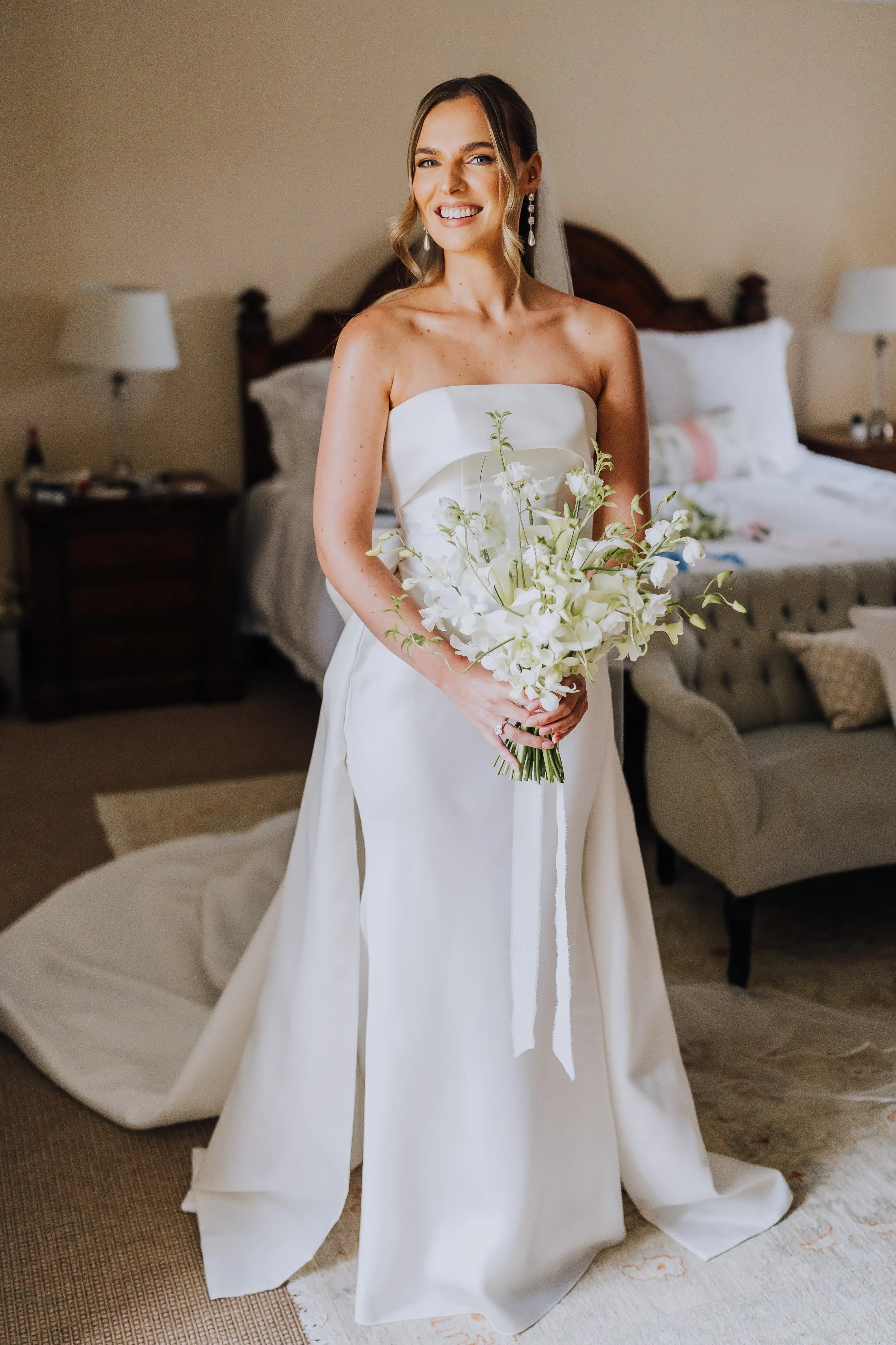 A smiling woman in a strapless white wedding dress holding a bouquet of white flowers, standing in a bedroom with a bed, lamps, and armchair in the background.