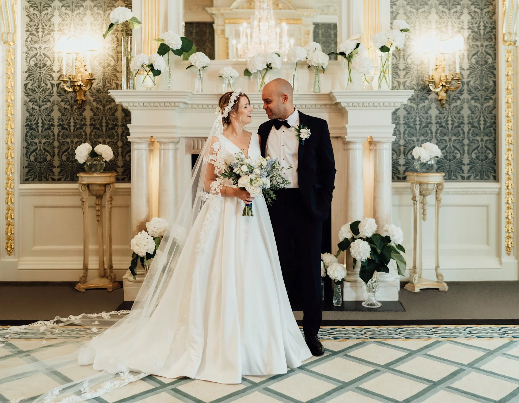 A bride and groom standing together in a decorated room, smiling and looking at each other, with white flowers in the background and on the fireplace.