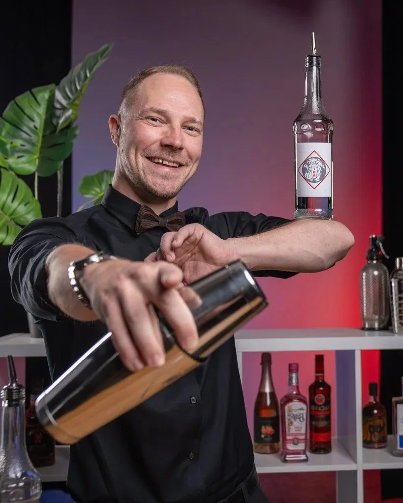 Mies nainen cocktail- tai drinkkimestar, jolla on yllään musta paita ja ruskea rusetti, valmistaa juomaa taikatemppuilun aikana, käyttämällä shakeria ja juoman pulloa, jonka yläpuolella on lasinen pullo, jossa on etiketti ja korkki, taustalla on hylly erilaisia alkoholipulloja ja kasvava viherkasvi.