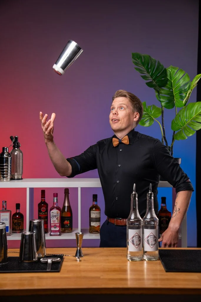Mies paloittelee metallista sekoitinta kokkailuun baaritiskillä, taustalla on alkoholipulloja ja viherkasvi, tausta on värikäs.