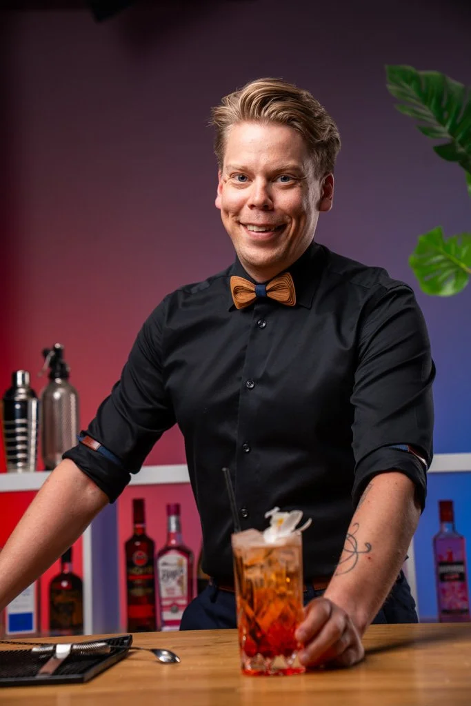 Hymyilevä baarimestari tarjoilee cocktailia baaripöydässä, taustalla pulloja ja kasvava viherkasvi.