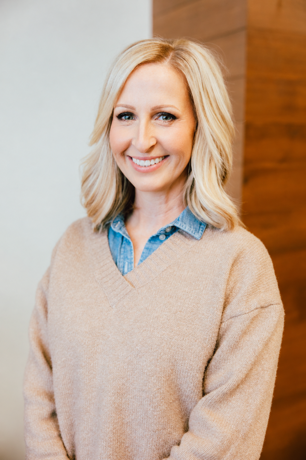 A woman with blonde hair and blue eyes is smiling at the camera, wearing glasses with braces, a tan sweater, and a blue collared shirt underneath, standing indoors.