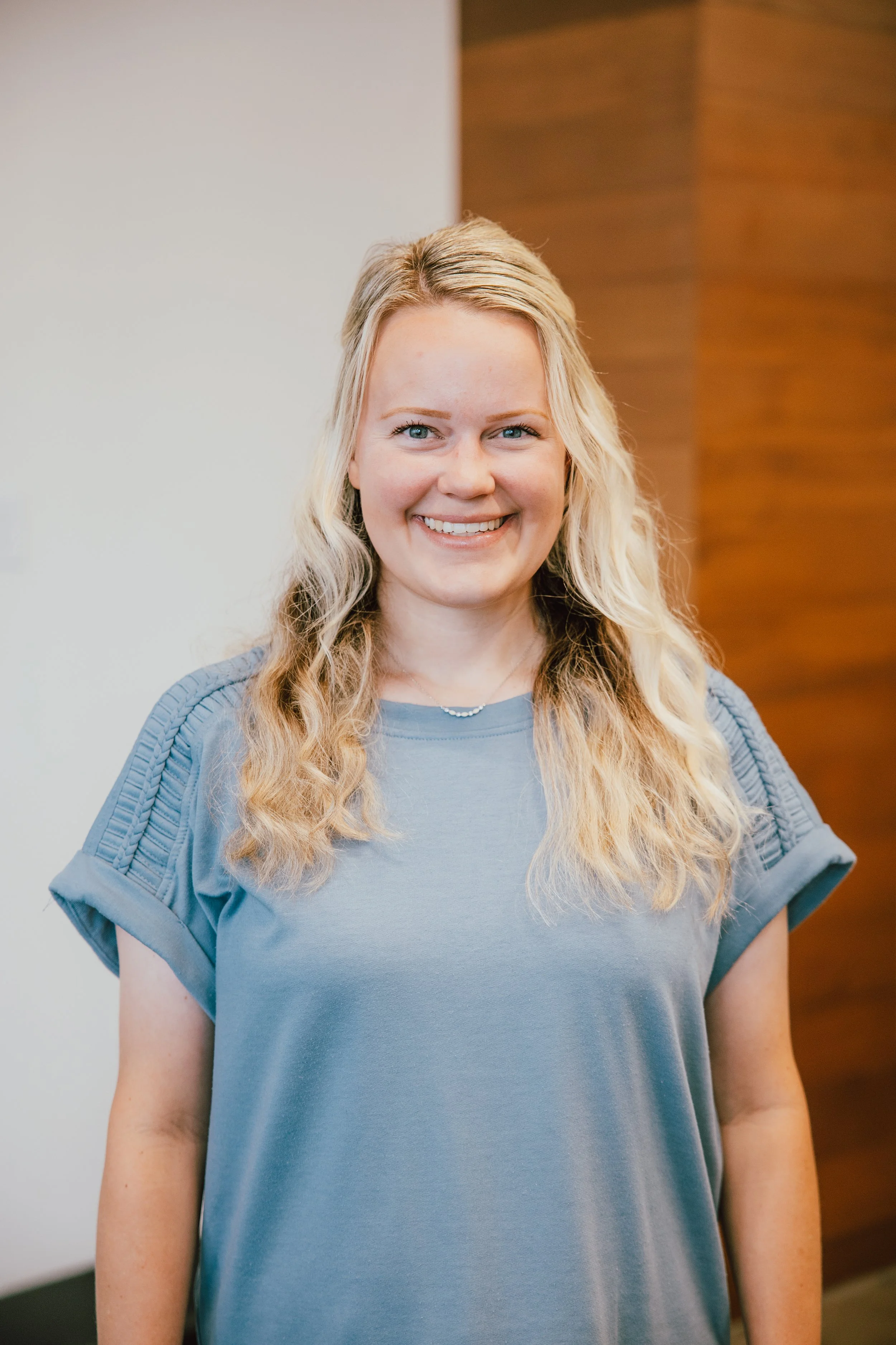 A woman with long wavy blonde hair, blue eyes, and a wide smile, wearing a light blue t-shirt with quilted shoulder details, standing indoors with a neutral wall and wood panel background.