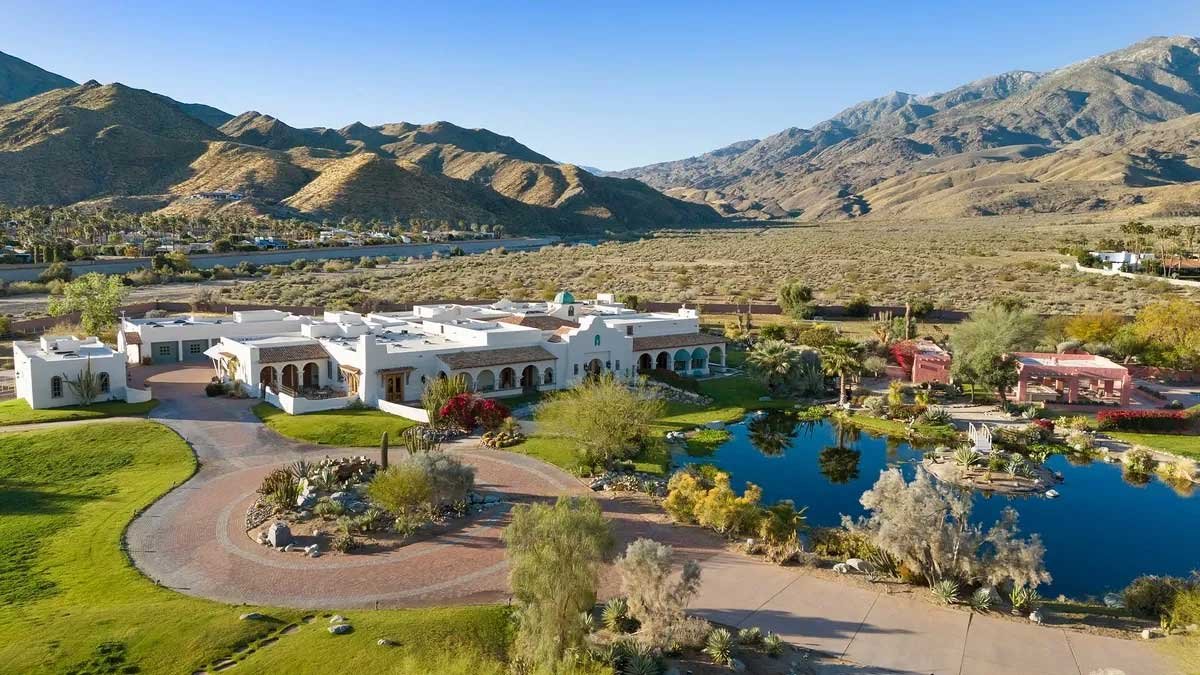 Aerial view of a large desert estate with adobe-style architecture, lush landscaping, palm trees, a small lake, and surrounding mountains.