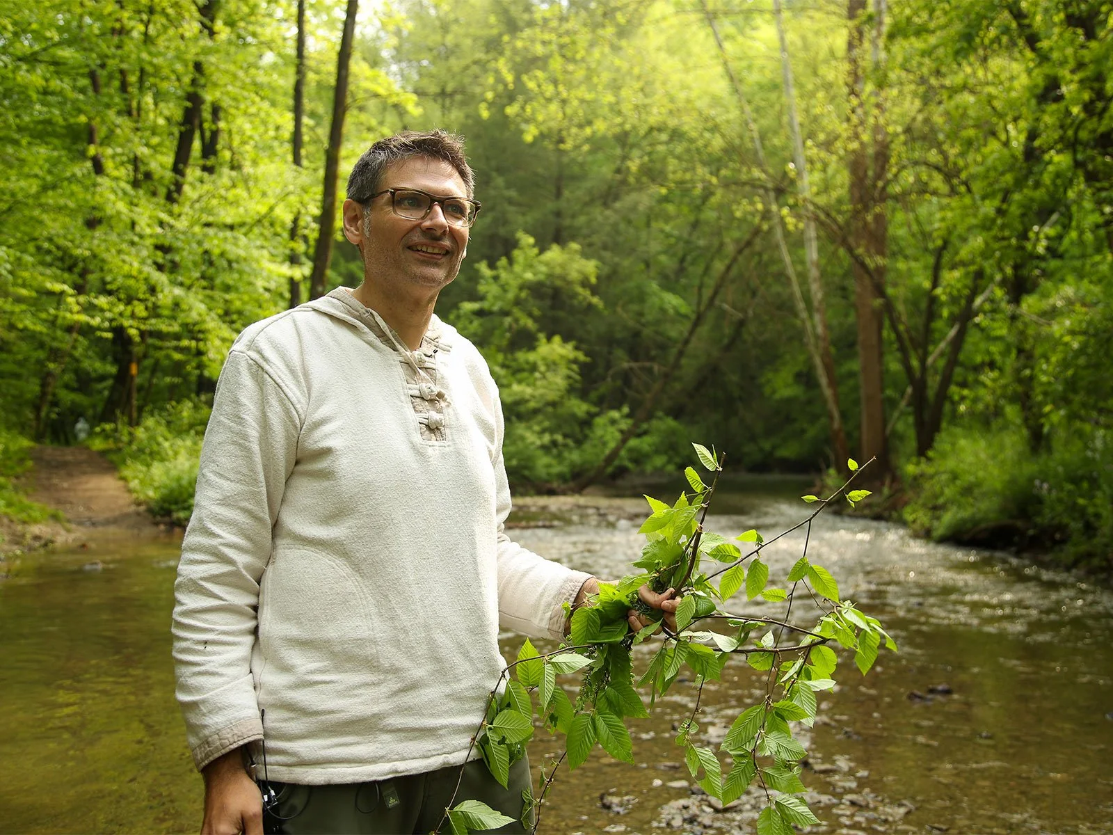 Man standing by a river in a forest, holding branches with green leaves, wearing a light-colored sweater.