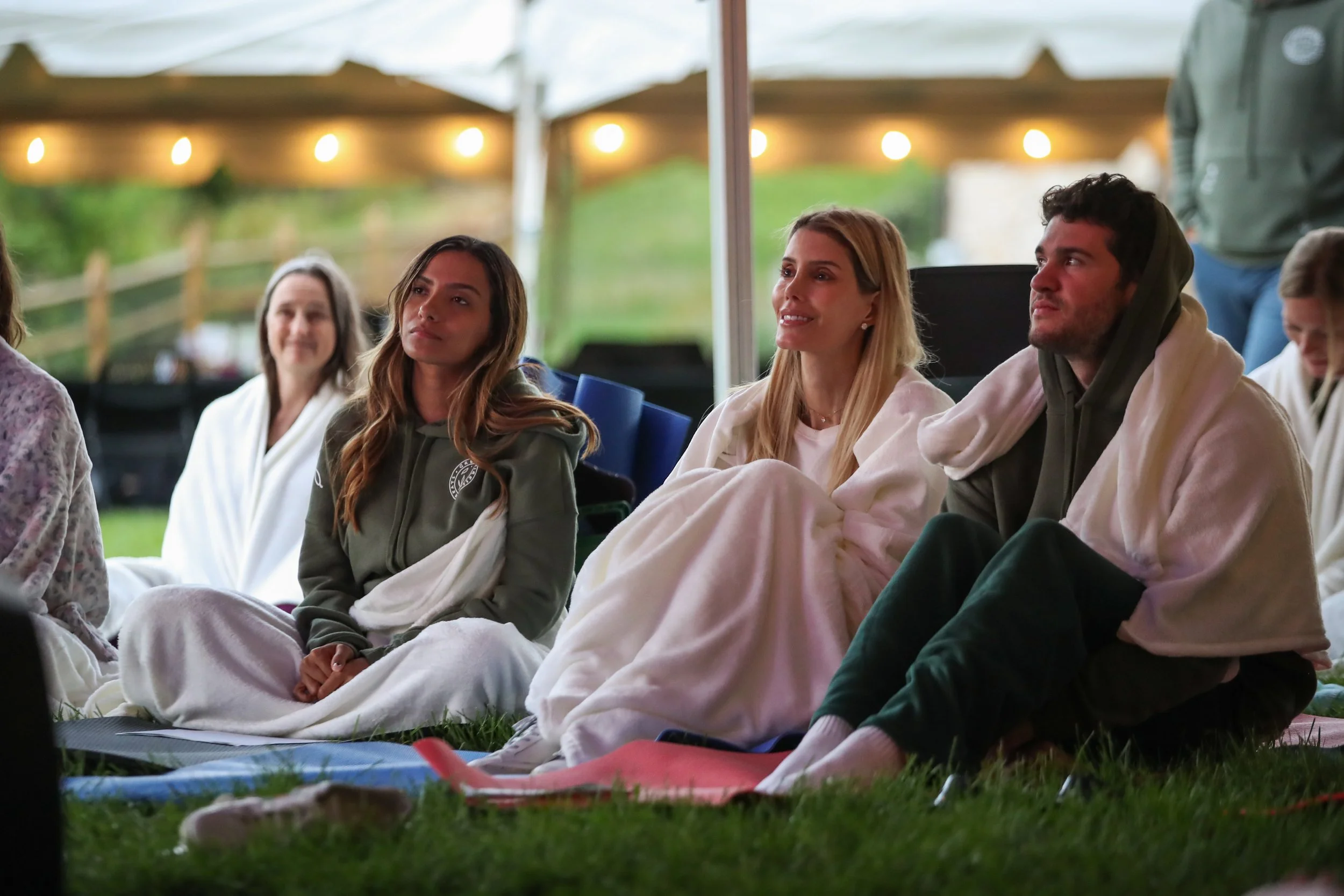 Group of people sitting outdoors on grass, wrapped in blankets, attending an event.