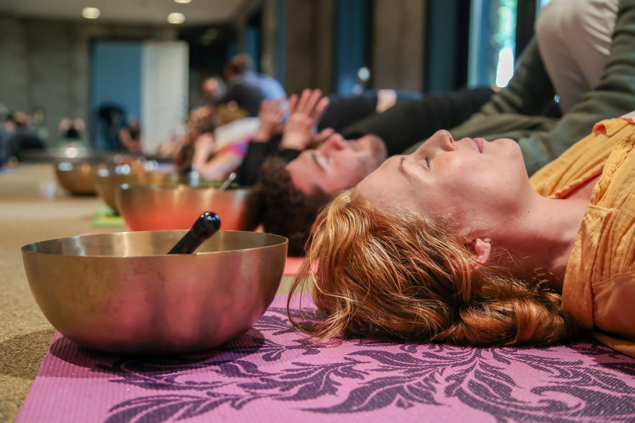 People lying down on yoga mats with Tibetan singing bowls beside them, participating in a sound meditation session.