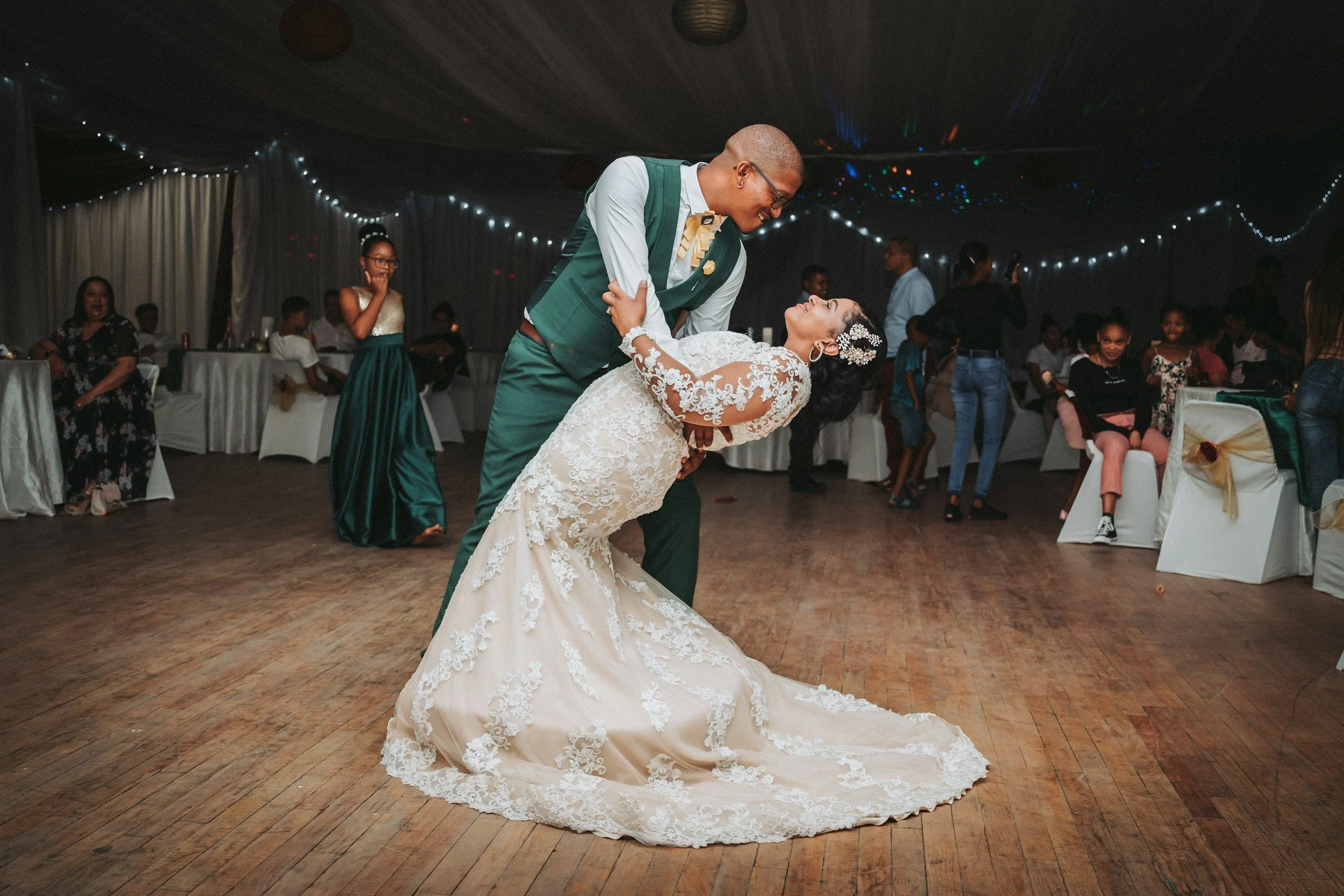 Bride and groom dancing at a wedding reception, the groom dipping the bride in a lace wedding gown; decorated venue with guests seated and standing, fairy lights in the background.